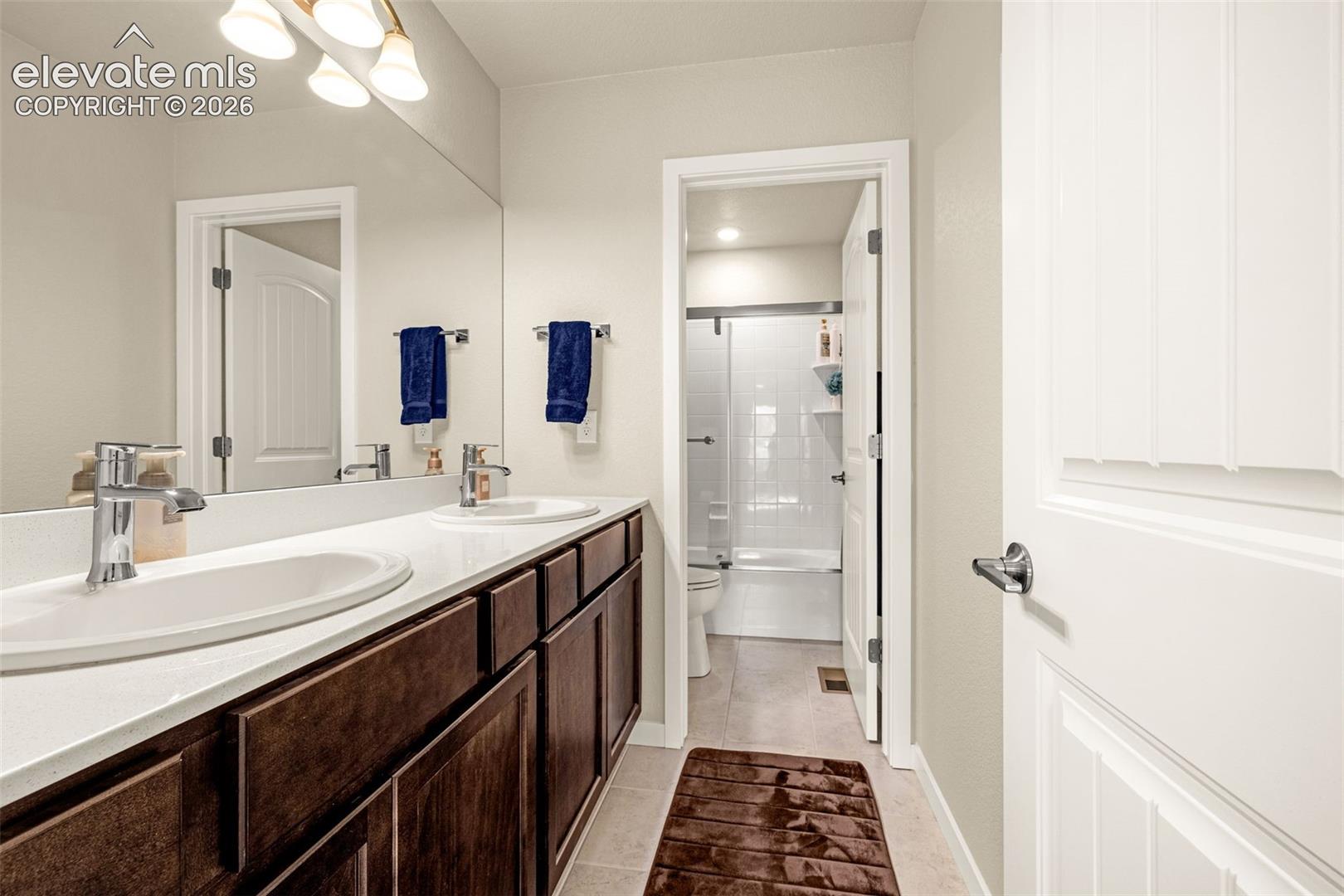 Bathroom featuring dark wood-finish cabinetry, dual white integrated sinks, a full-width vanity mirror, and polished metal hardware