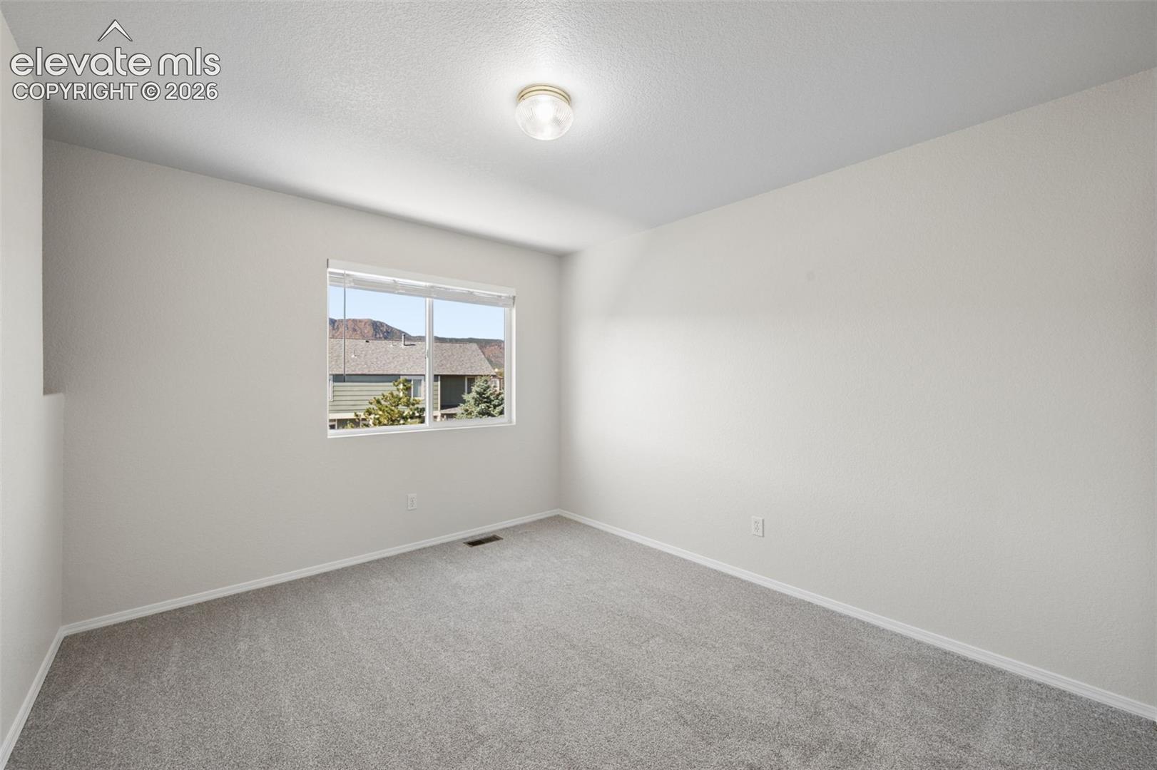 Neutral-toned interior space featuring light-colored wall paint, plush carpeting, a centered ceiling-mounted light fixture, and a window with a mountain backdrop