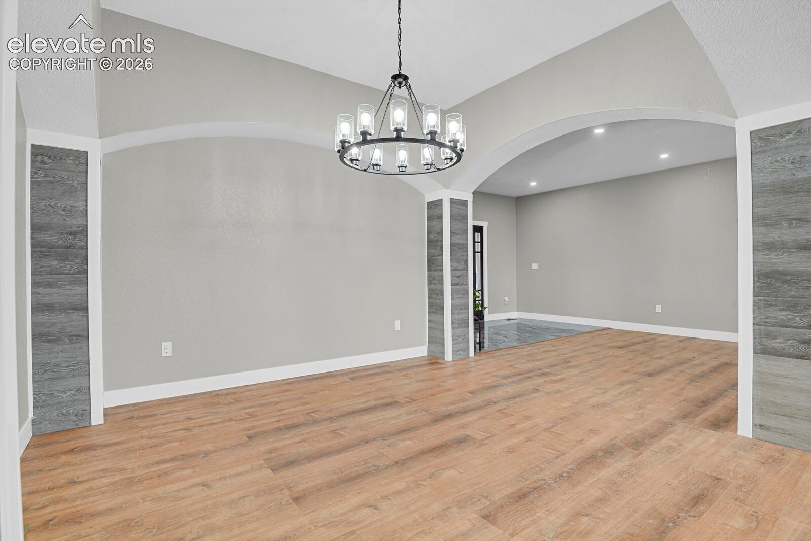 Open-concept interior featuring vaulted ceilings, wood-finish flooring, a multi-light chandelier, and decorative wood-panel accent columns framing a large arched opening