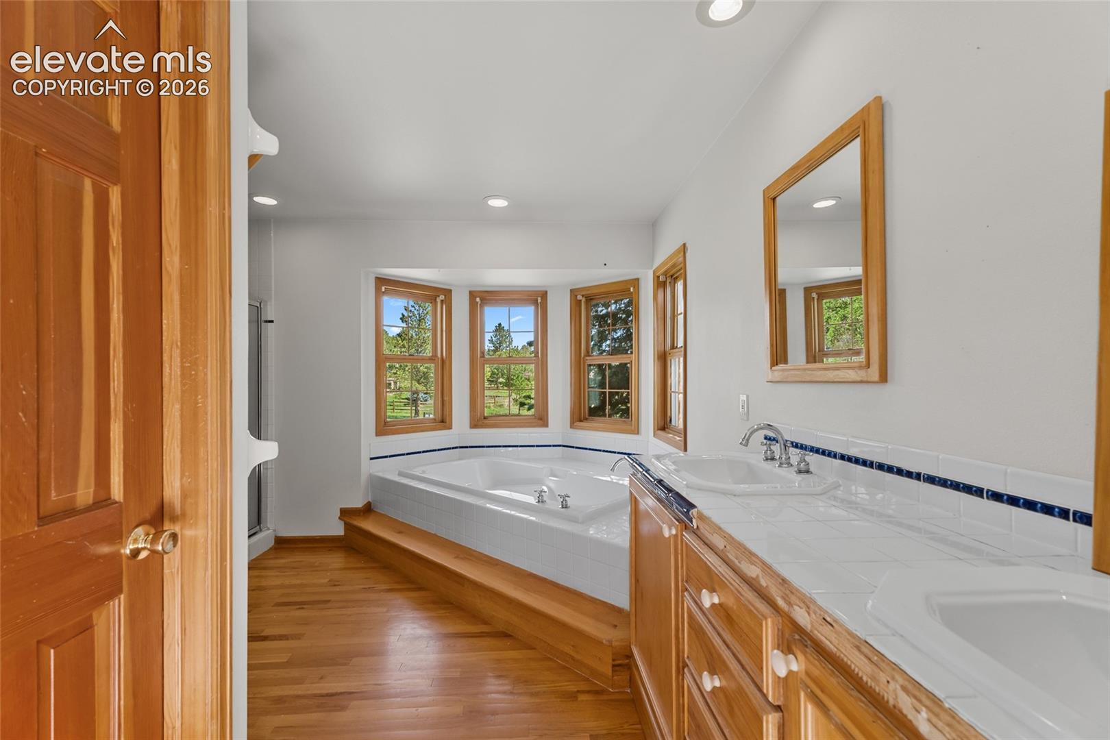 Hardwood-finish flooring, built-in soaking tub with tiered wood-finish surround, dual vanity with tile countertop, and multiple windows with wood-finish trim