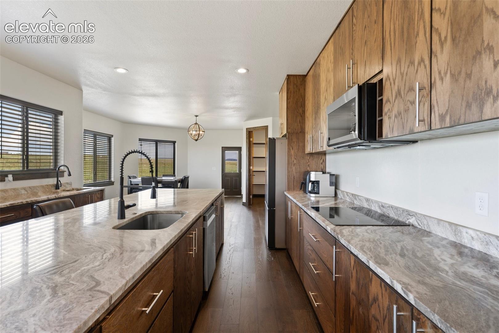 Open-concept kitchen featuring an expansive island with an undermount sink, stone countertops, wood-finish cabinetry, and integrated stainless steel appliances