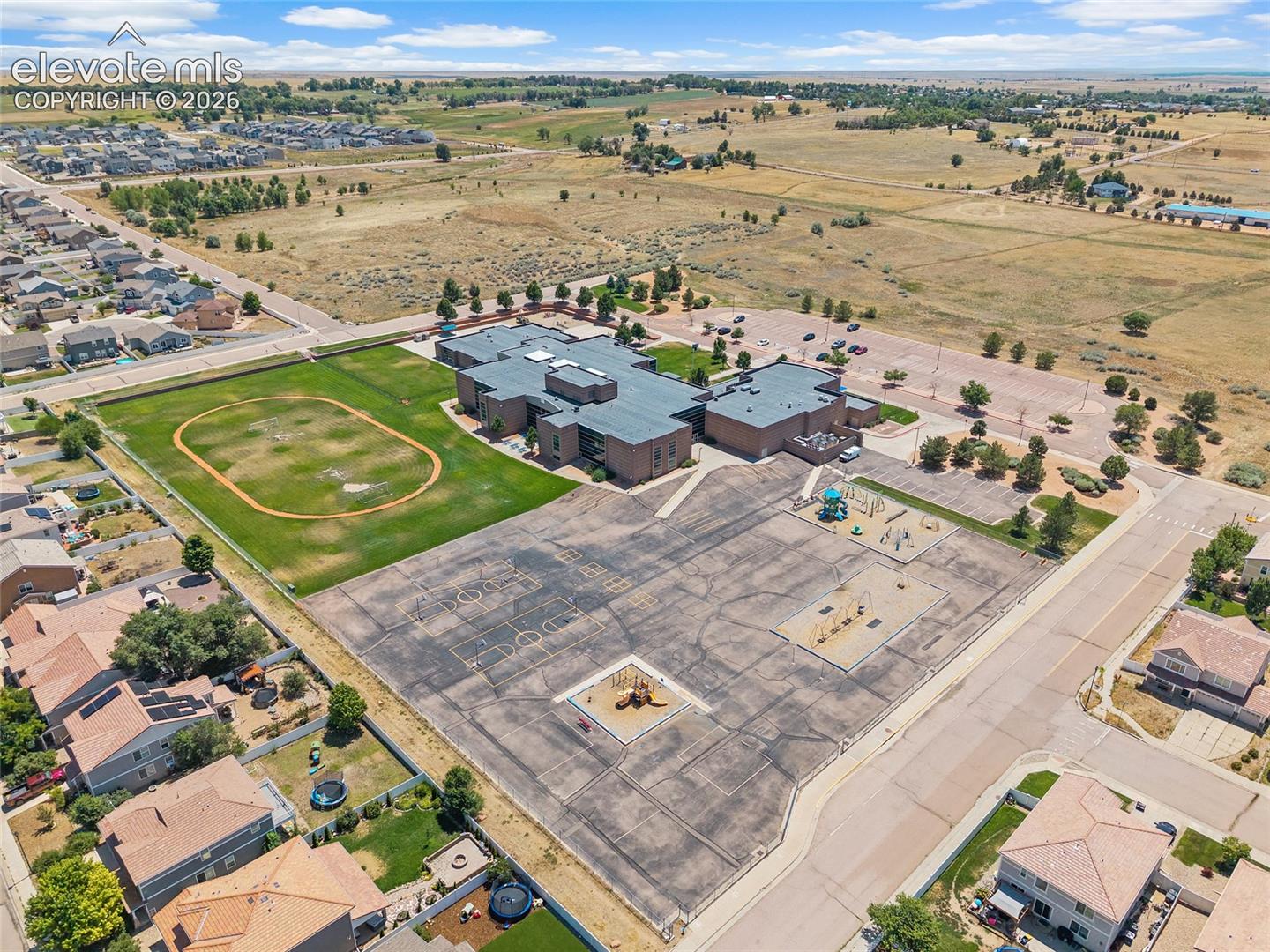 Aerial perspective featuring a multi-wing brick building, an expansive grass field, a paved play area, and integrated parking lots