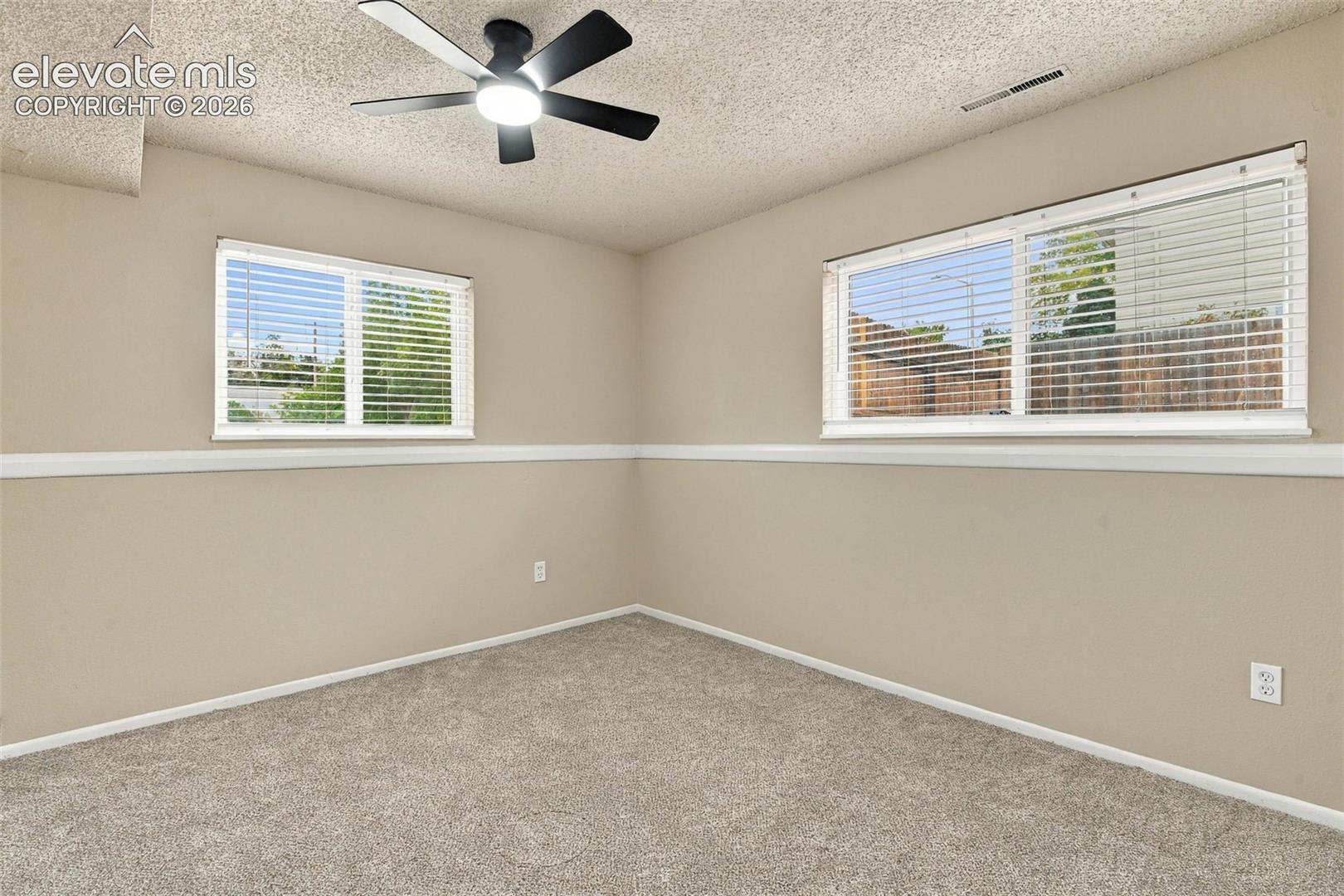 Neutral-toned room featuring textured ceiling, carpeted flooring, white chair rail molding, dual windows with fitted blinds, and a black multi-blade ceiling fan with integrated light