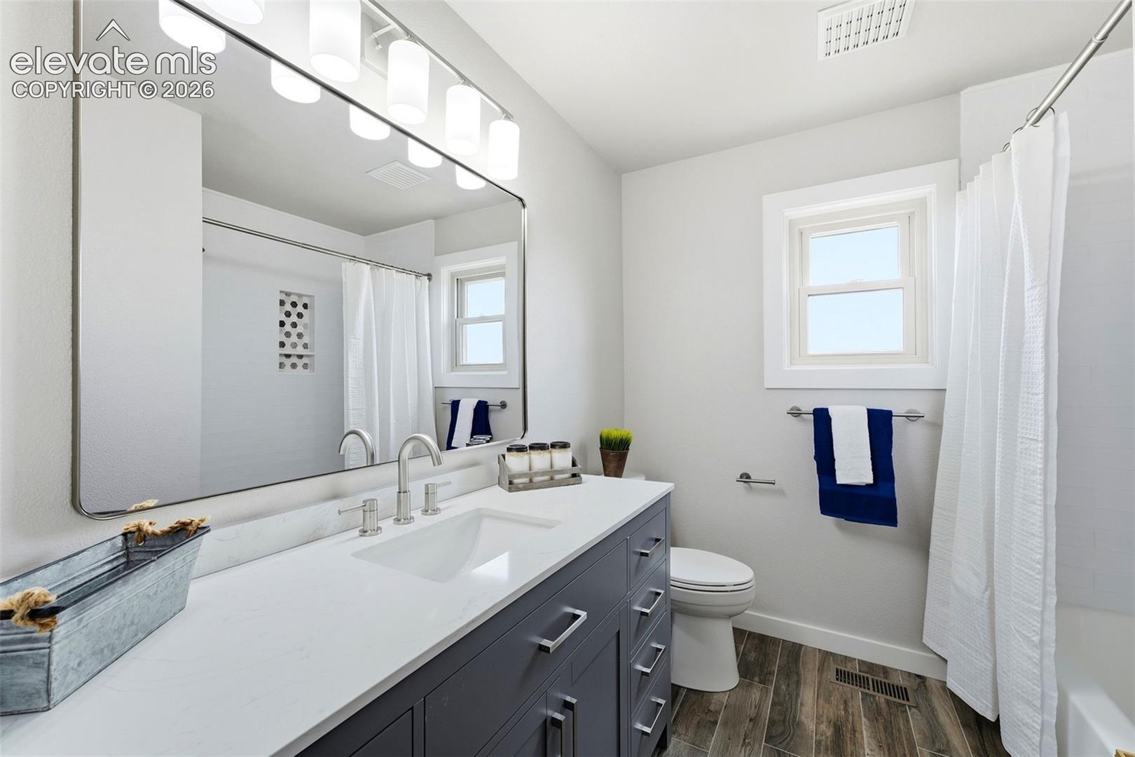 White vanity with a rectangular sink, wall-mounted mirror, brushed nickel fixtures, and wood-finish flooring