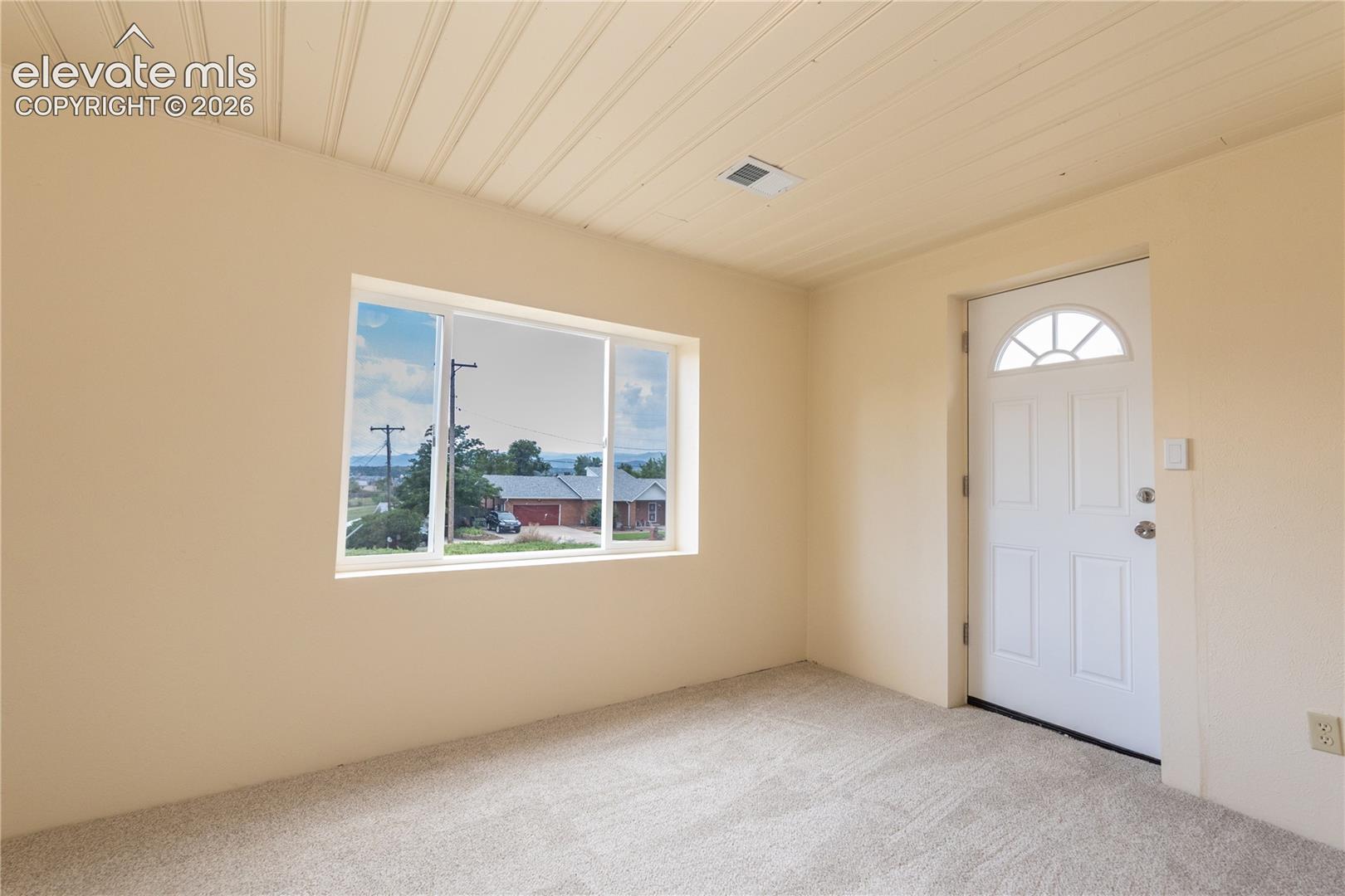 Interior space featuring neutral wall tones, carpeted flooring, a paneled ceiling, a wide picture window, and an exterior door with a semi-circular transom window
