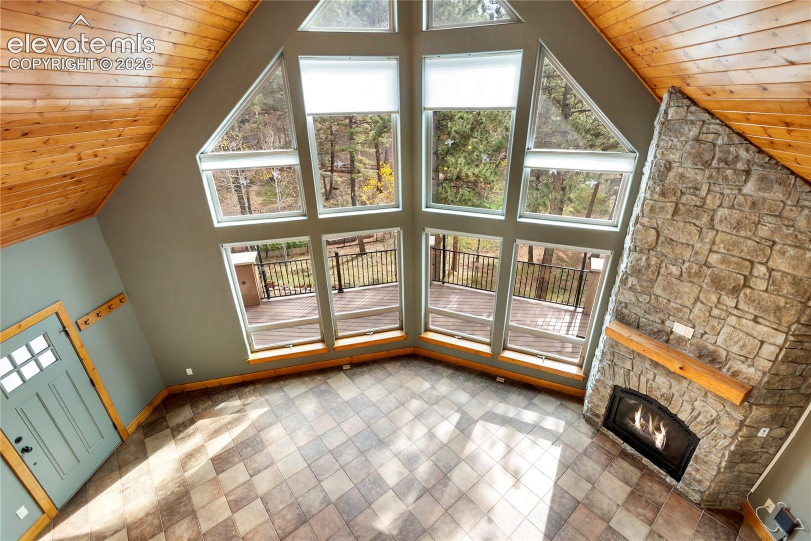 Vaulted wood-plank ceiling, multi-pane window wall, full-height stone masonry fireplace, tiled flooring, and wood-trimmed entry door