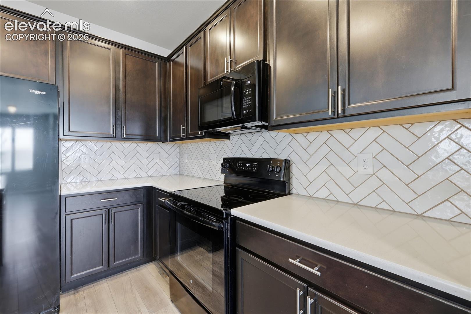 Modern kitchen featuring dark wood cabinetry, white herringbone tile backsplash, light-toned countertops, and black appliances including a refrigerator and range with an overhead microwave