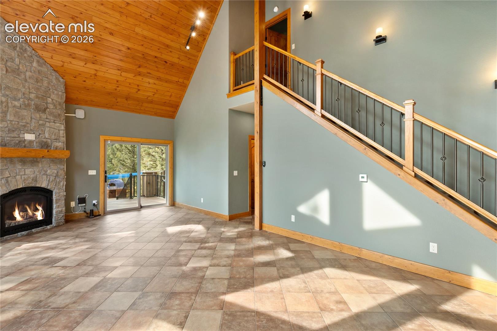 Vaulted ceiling with exposed wood paneling, stone-faced fireplace with wood mantel, tile flooring, and staircase featuring wooden handrails with metal balusters