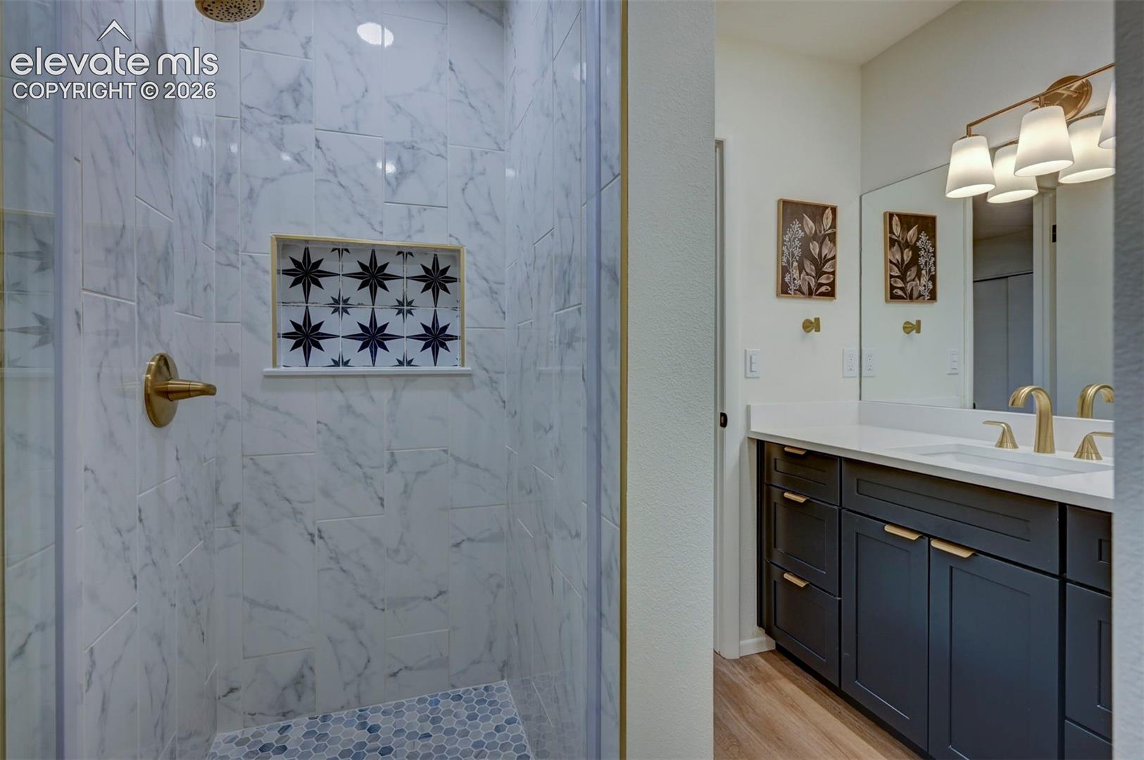 Shower with stone-finish tile surround and decorative inset, brass-tone hardware, vanity with solid-surface countertop, dark-toned cabinetry, and multi-light fixture