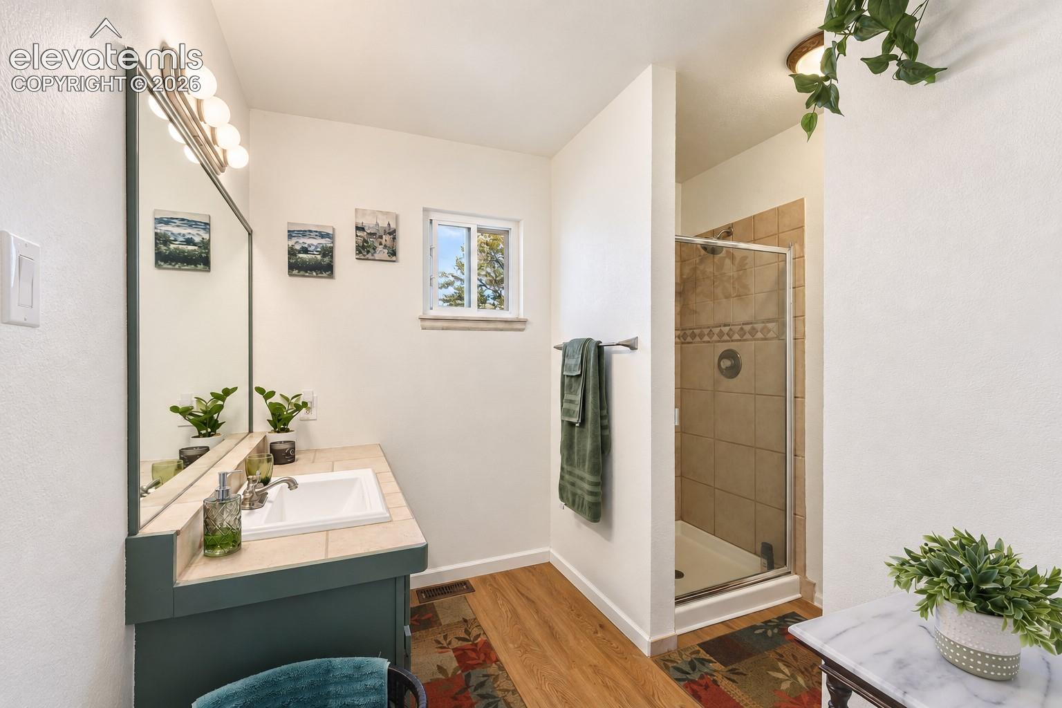 Bathroom featuring a vanity with tile-surfaced countertop, integrated sink, framed wall mirror, wood-finish flooring, and a tiled shower enclosure with a glass door