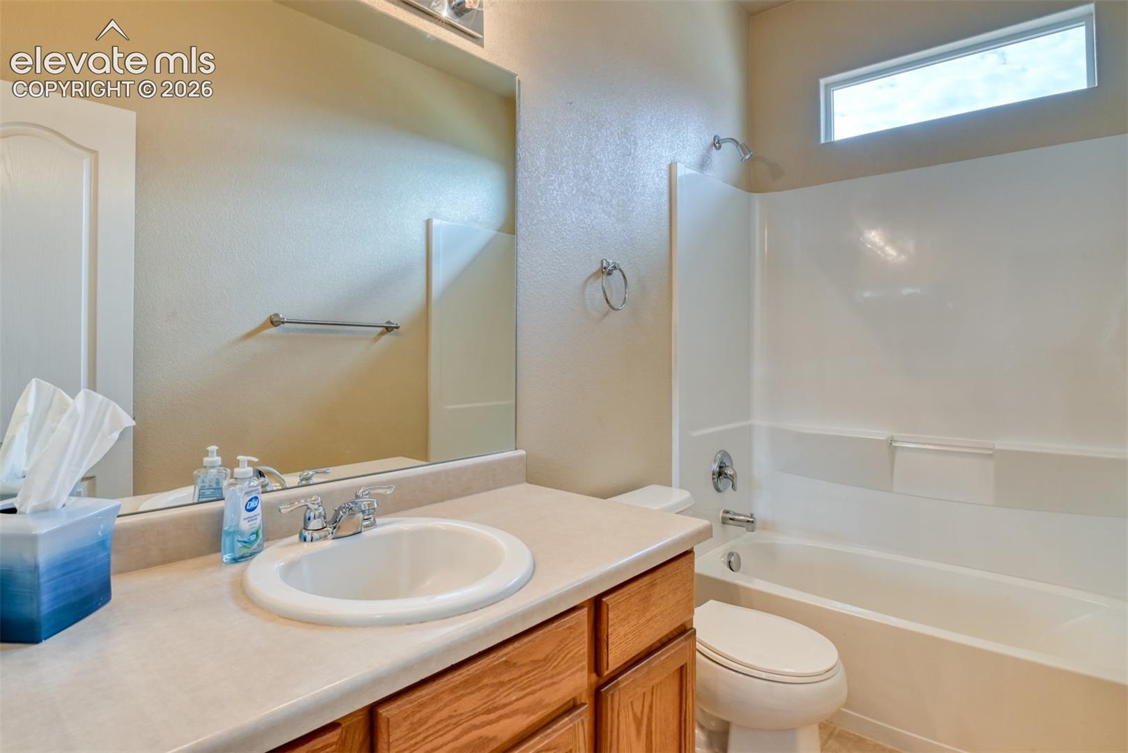 Bathroom featuring a single-sink vanity with solid-surface countertop and wood-finish cabinetry, full-width vanity mirror, and a combination tub and shower enclosure with transom window