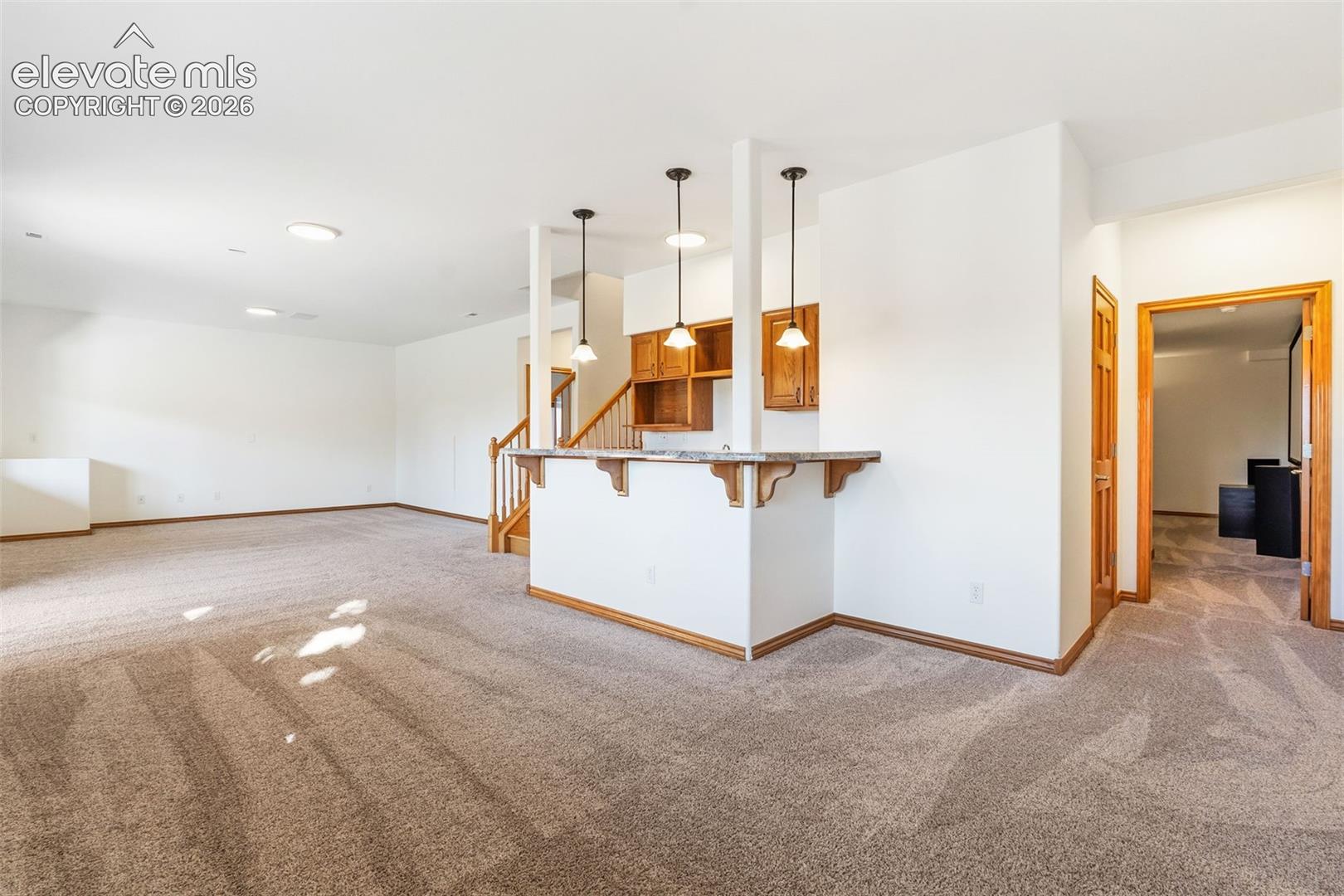 Open living area featuring neutral wall-to-wall carpeting, a built-in breakfast bar with stone-finish countertop, three pendant light fixtures, and wood-trim cabinetry