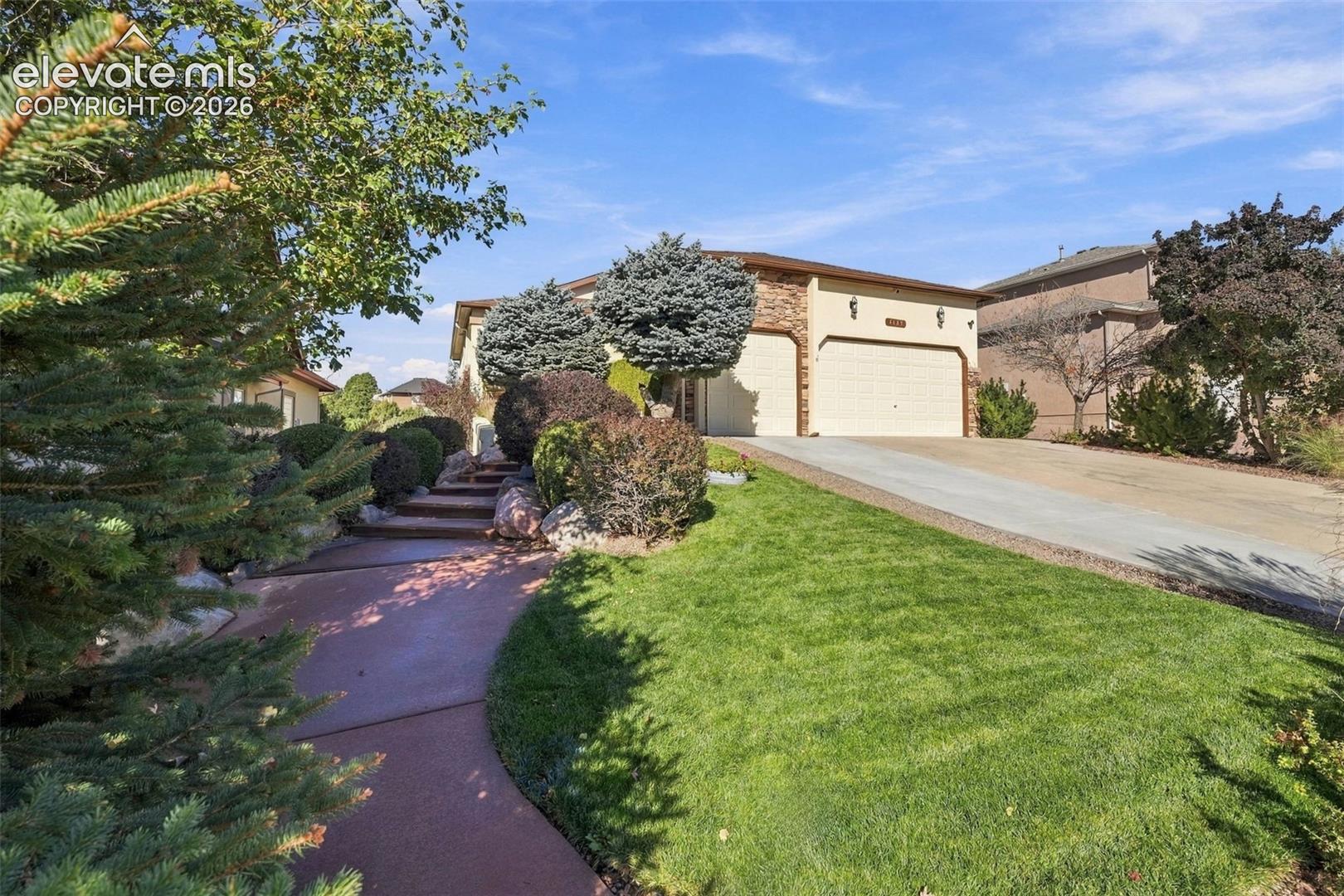 Stucco exterior featuring a dual-bay garage, stone masonry accents, a paved walkway, and a landscaped front lawn with mature shrubbery