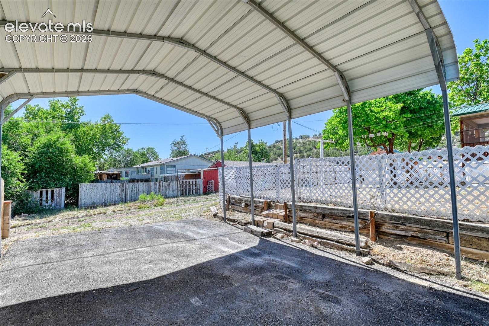 Metal-roofed carport with asphalt parking surface, white lattice fencing, and tiered timber retaining wall