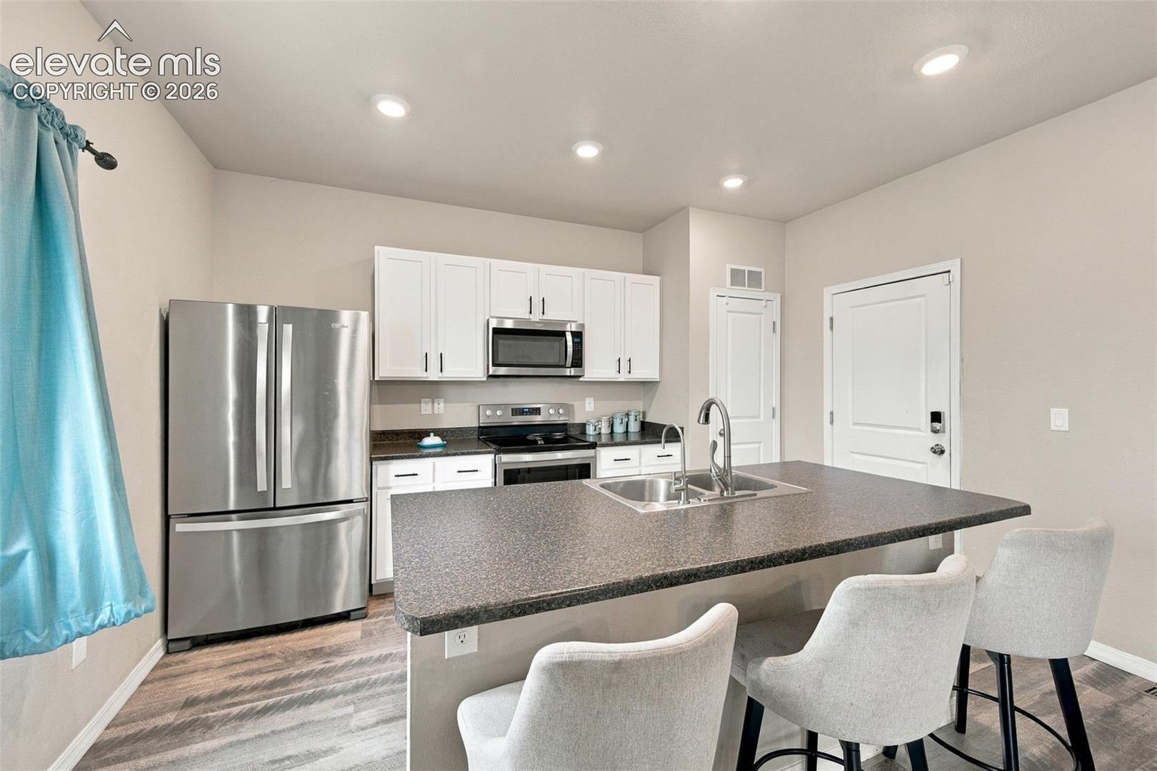 Kitchen featuring white shaker-style cabinetry, stainless steel appliances, a central island with a dual-basin sink, and wood-finish flooring