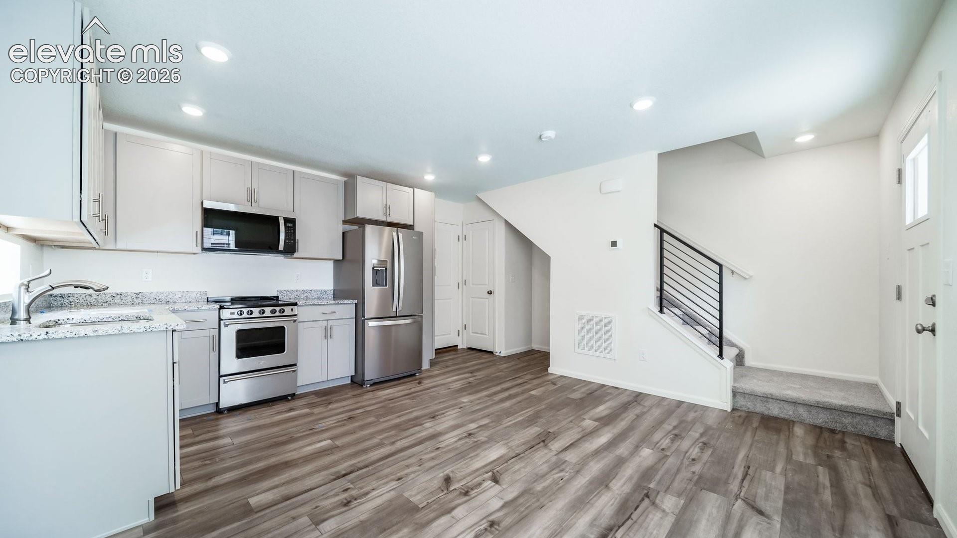 Kitchen featuring light-toned cabinetry, stone-finish countertops, stainless steel appliances, wood-finish flooring, and a staircase with a horizontal metal railing
*photos of a model home*