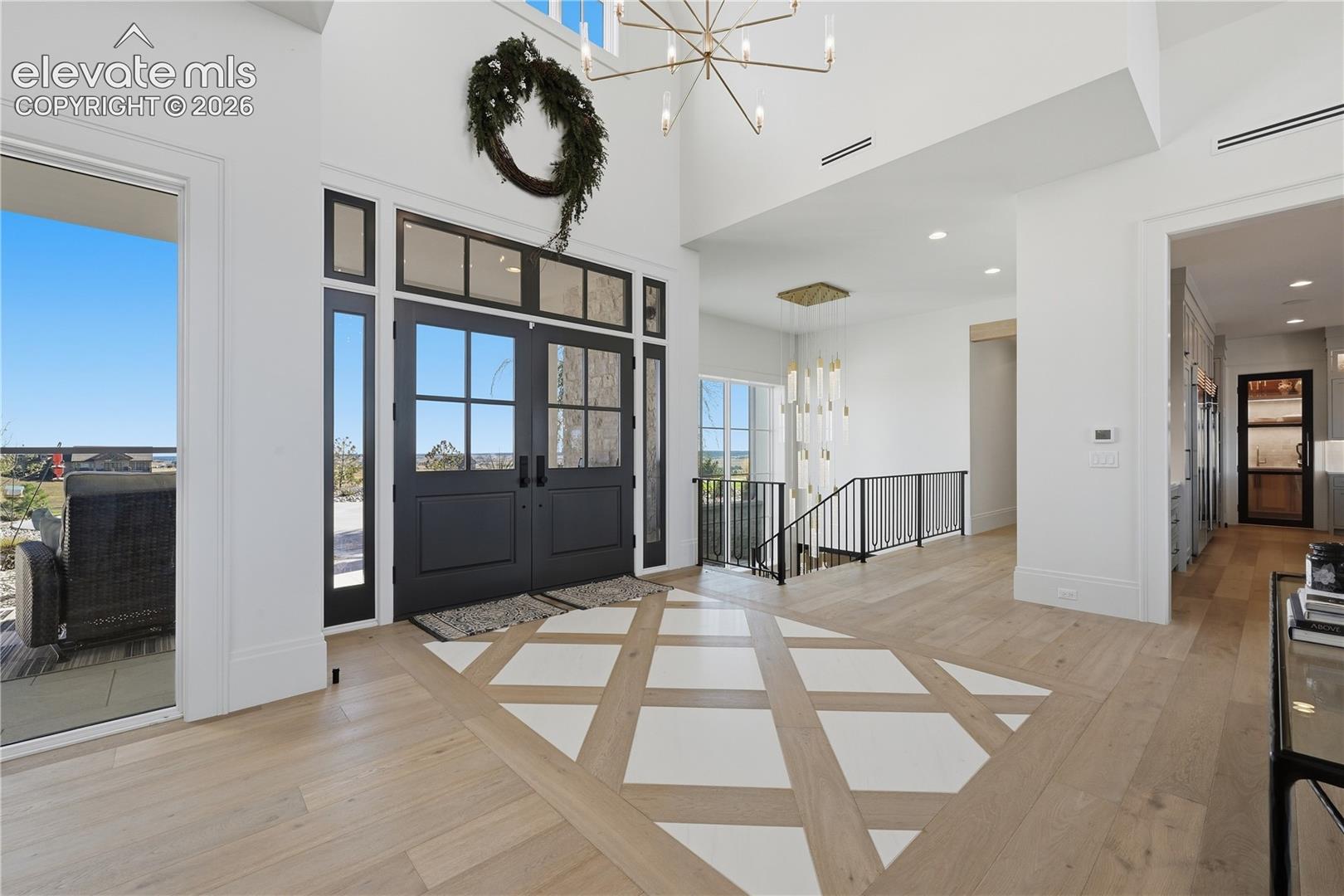 Grand foyer featuring high ceilings, double entry doors with glass panes, wood-finish flooring with a decorative geometric inlay, a multi-tiered modern chandelier, and a metal-railed staircase