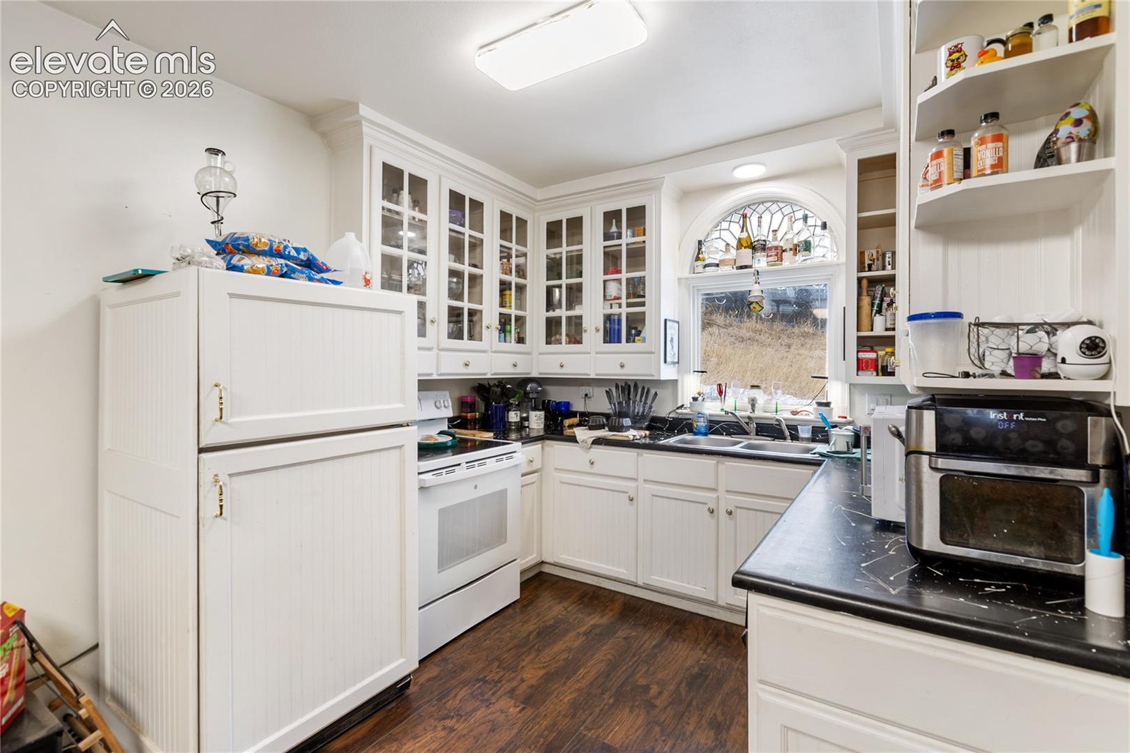 Kitchen featuring white cabinetry with glass-front uppers, beadboard paneling, dark countertops, wood-finish flooring, and an arched window