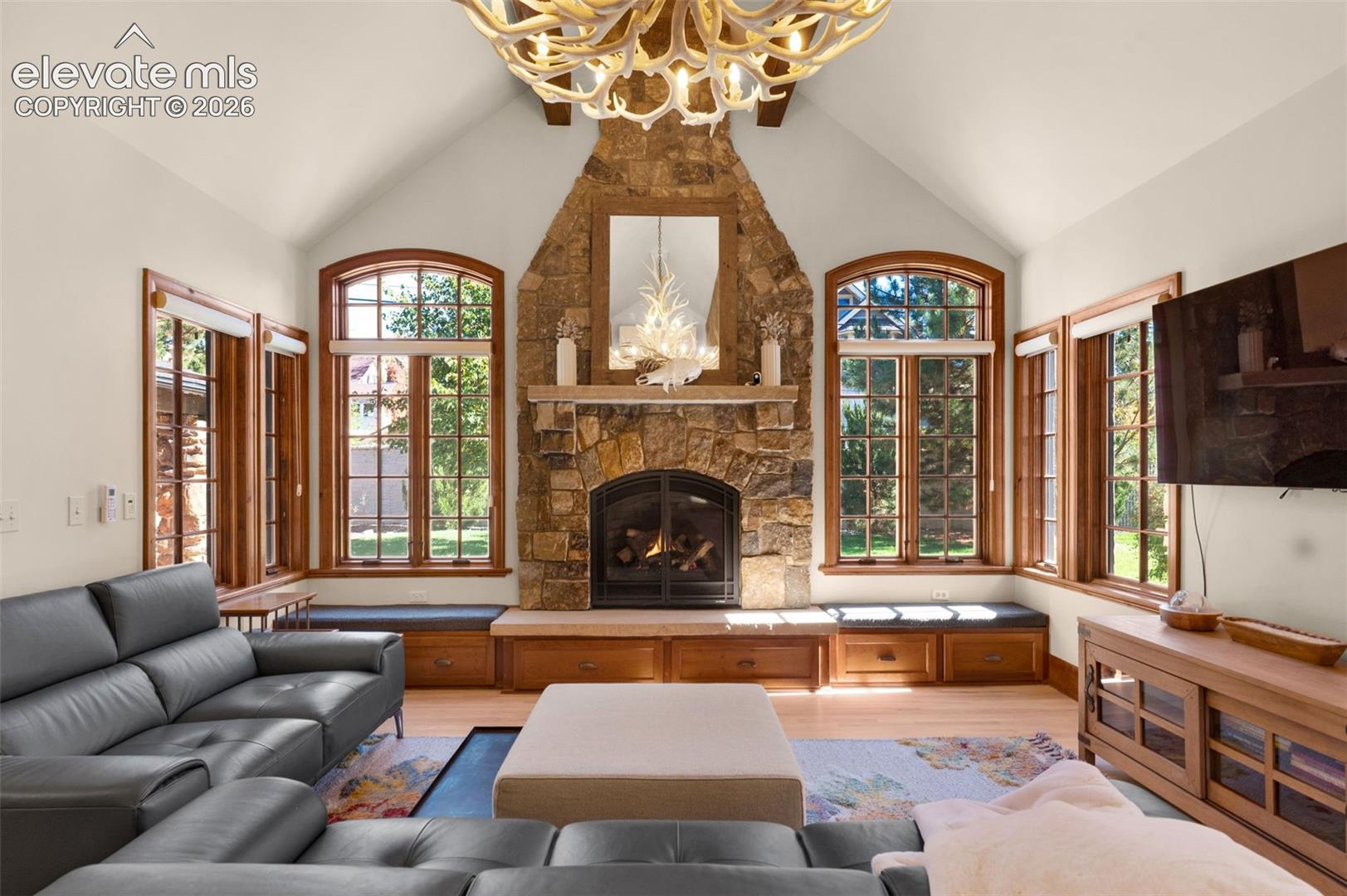 Vaulted ceilings with exposed timber beams, floor-to-ceiling stone fireplace with a substantial mantel, built-in window seating with integrated cabinetry, and arched multi-pane windows
