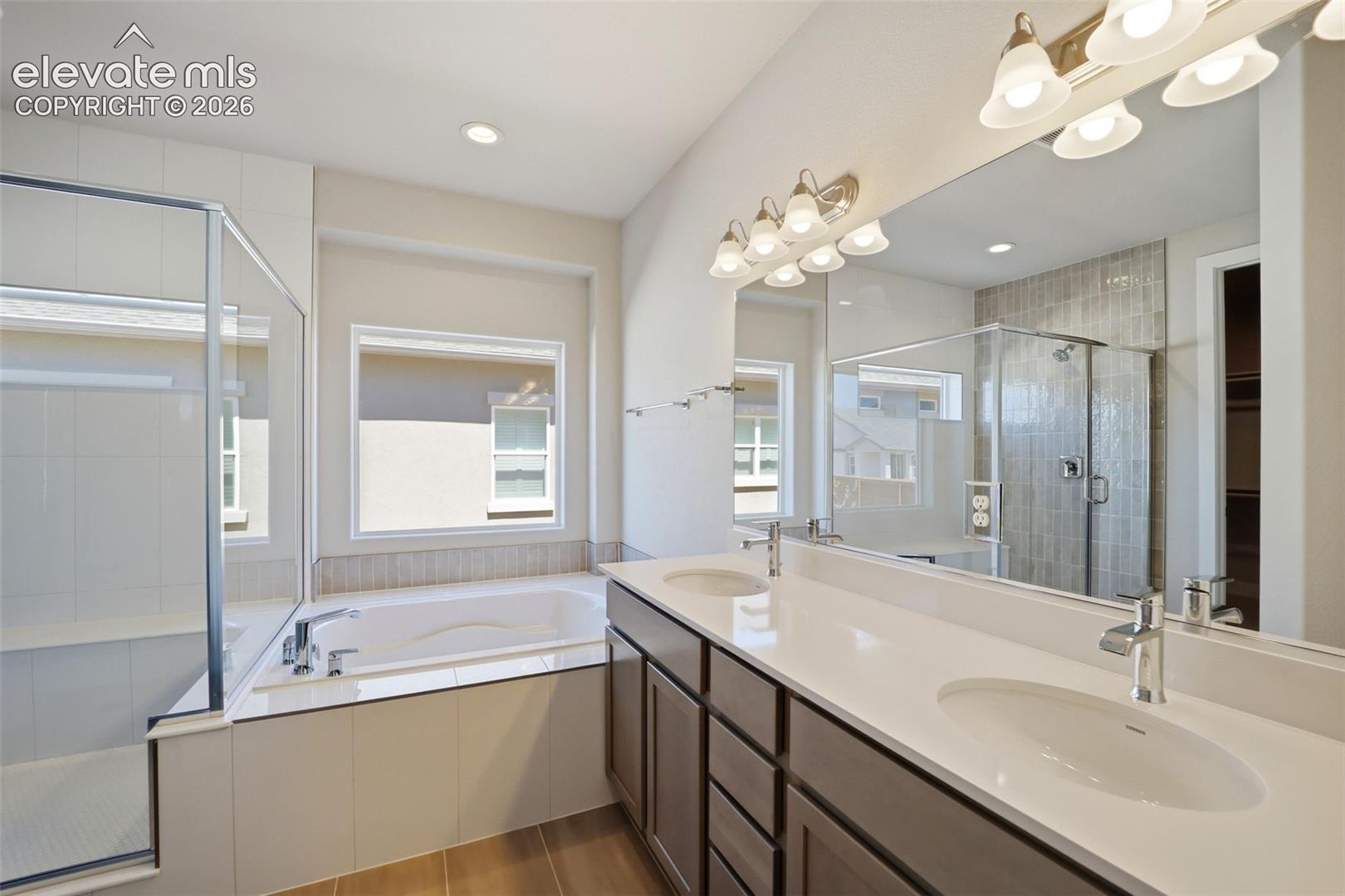 Double vanity with solid surface countertops, drop-in sinks, and full-width wall mirror, accompanied by a soaking tub and glass-enclosed shower with tile surround