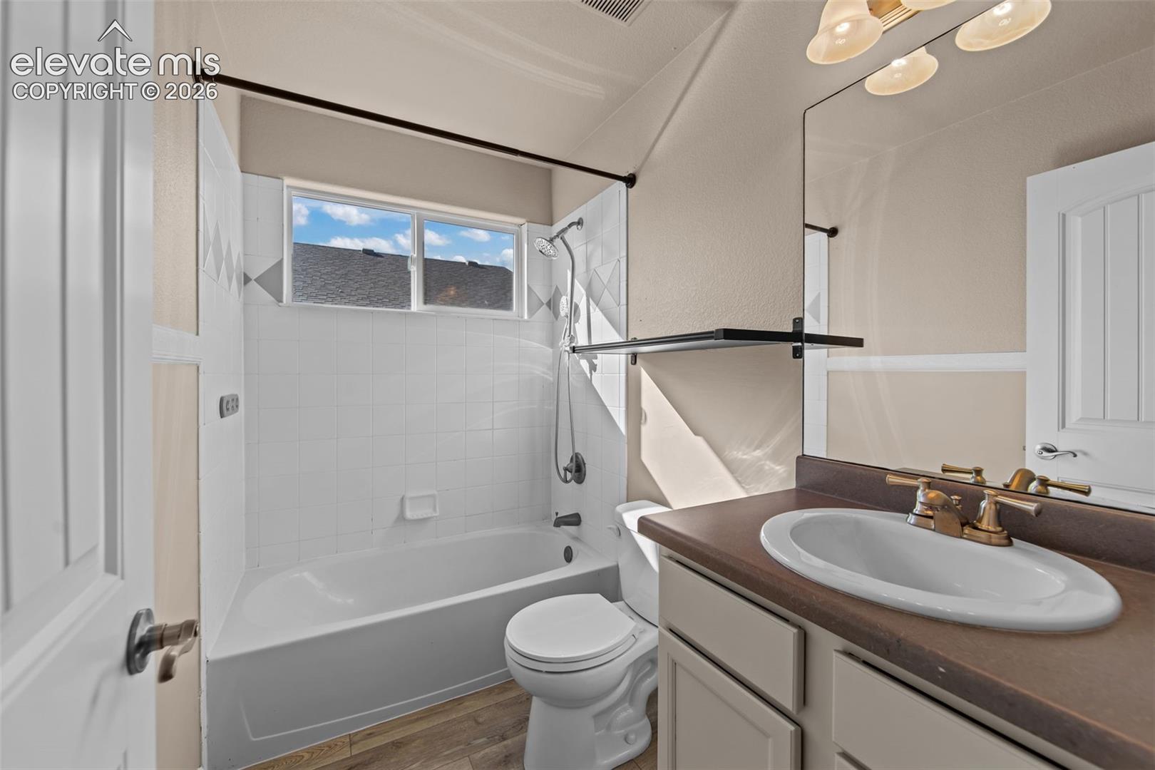 Bathroom featuring a composite countertop with an integrated oval sink, brass-finish faucet, wood-finish flooring, and a shower-tub combination with white wall tile