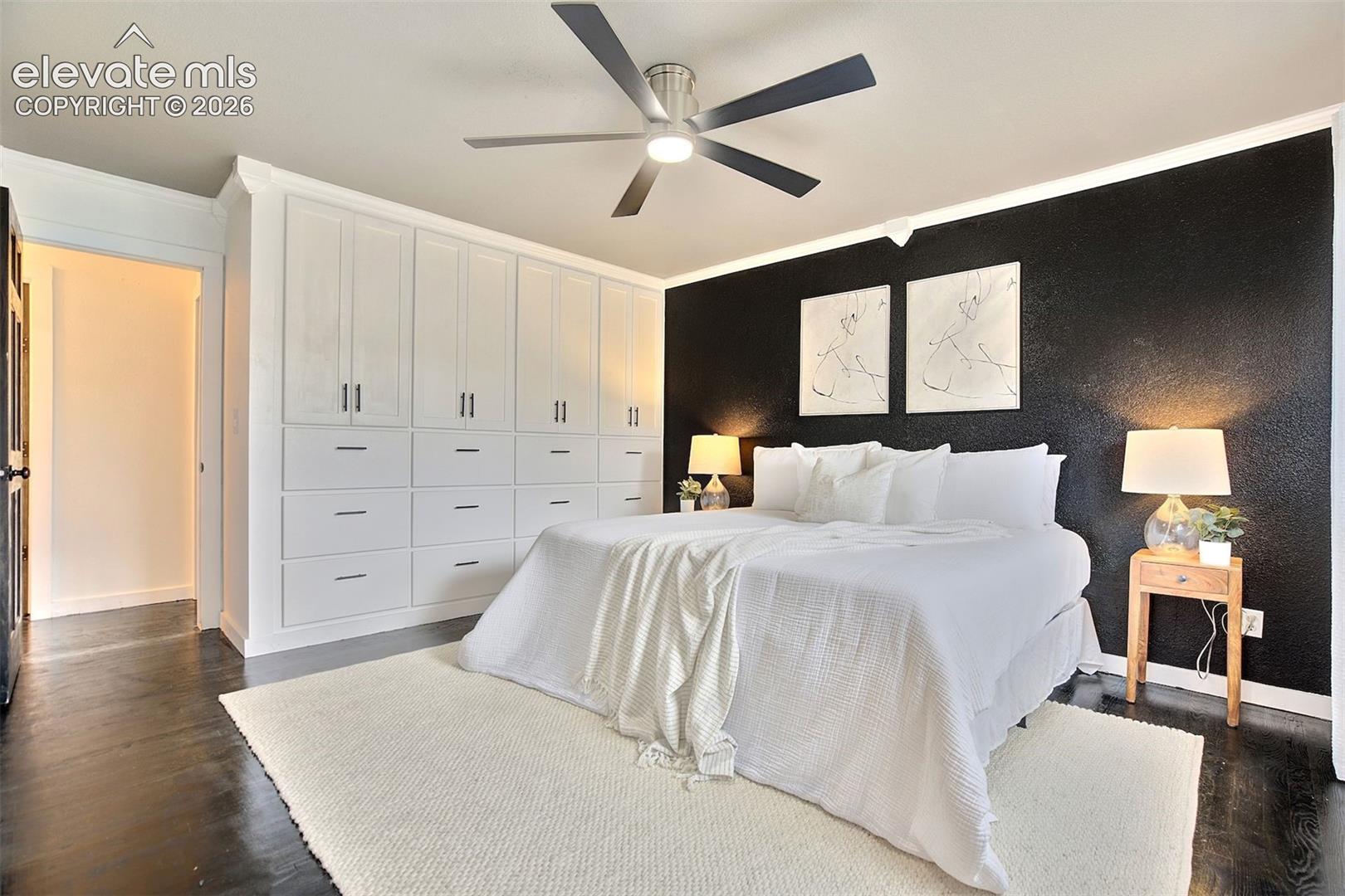 Extensive wall-to-wall white built-in cabinetry, dark wood-finish flooring, accent wall, and a multi-blade ceiling fan with integrated lighting