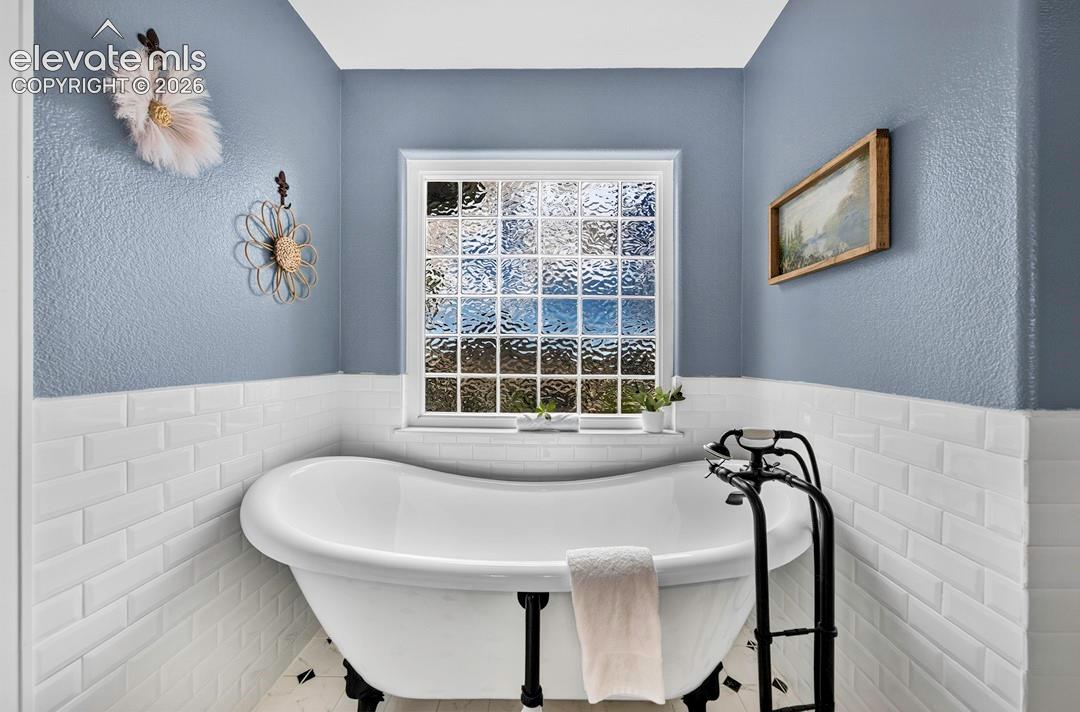 Freestanding white soaking tub with matte black hardware, white subway tile wainscoting, and a window featuring decorative textured glass