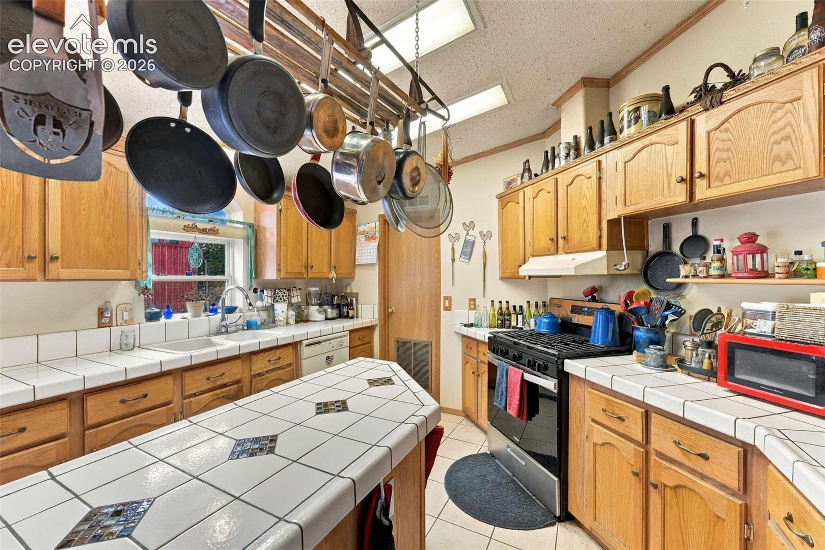 Kitchen featuring wood cabinetry, tiled countertops with decorative inlays, a central island, a hanging pot rack, and built-in range
