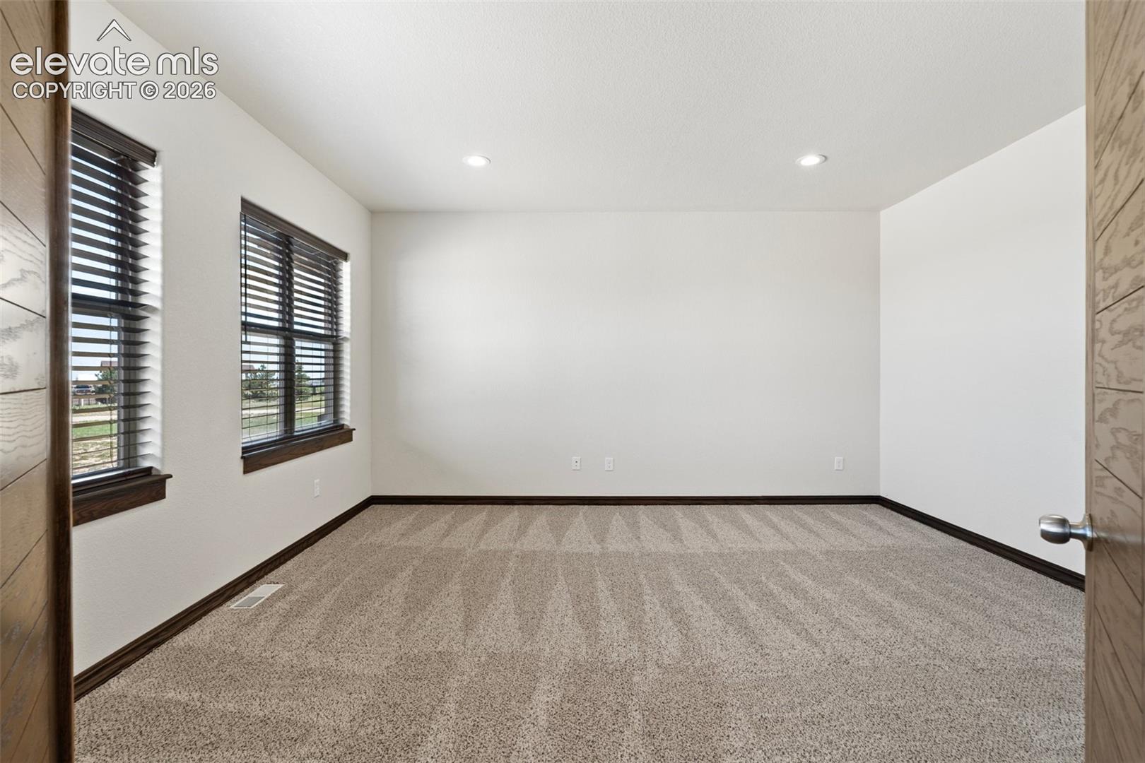 Spacious office featuring neutral carpeting, recessed ceiling lighting, two windows with dark wood blinds and trim, and a modern wood-paneled door