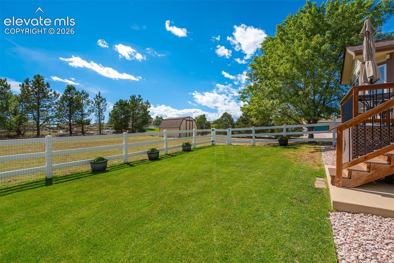 Expansive backyard featuring a manicured lawn, white post-and-rail fencing, a detached storage structure, and a wood-framed deck