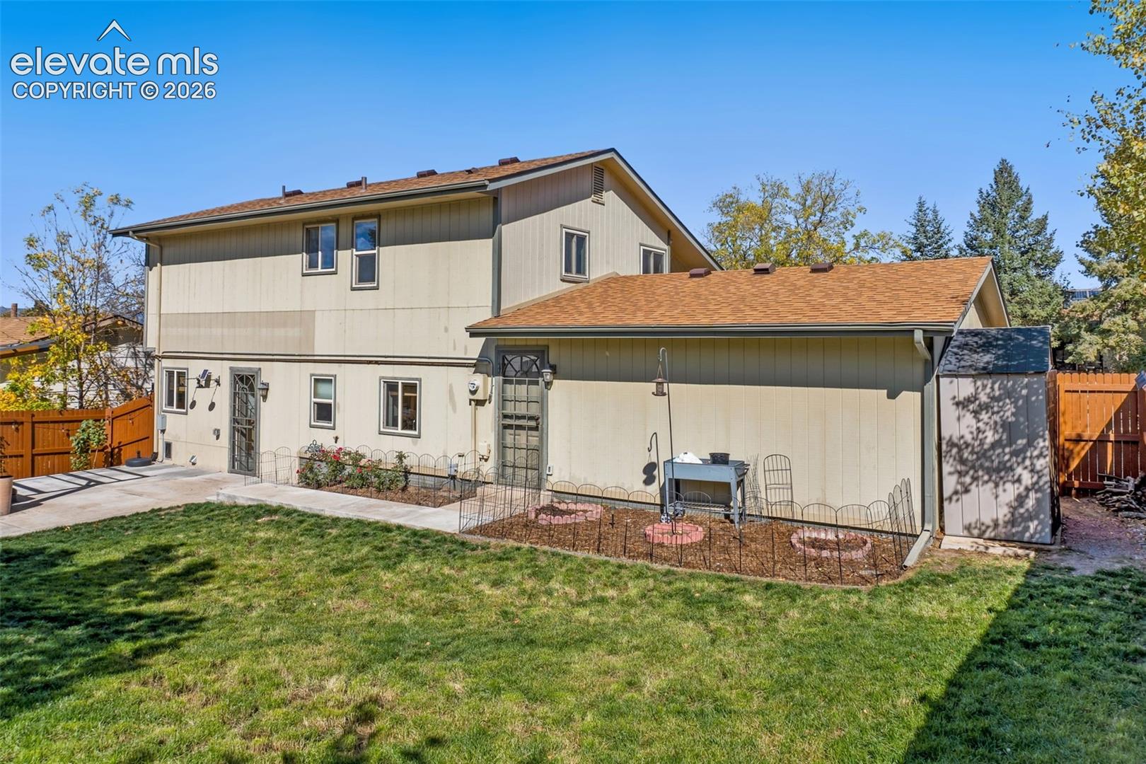 Exterior rear elevation featuring vertical siding, shingle roof, concrete patio, perimeter wood fencing, and designated garden beds