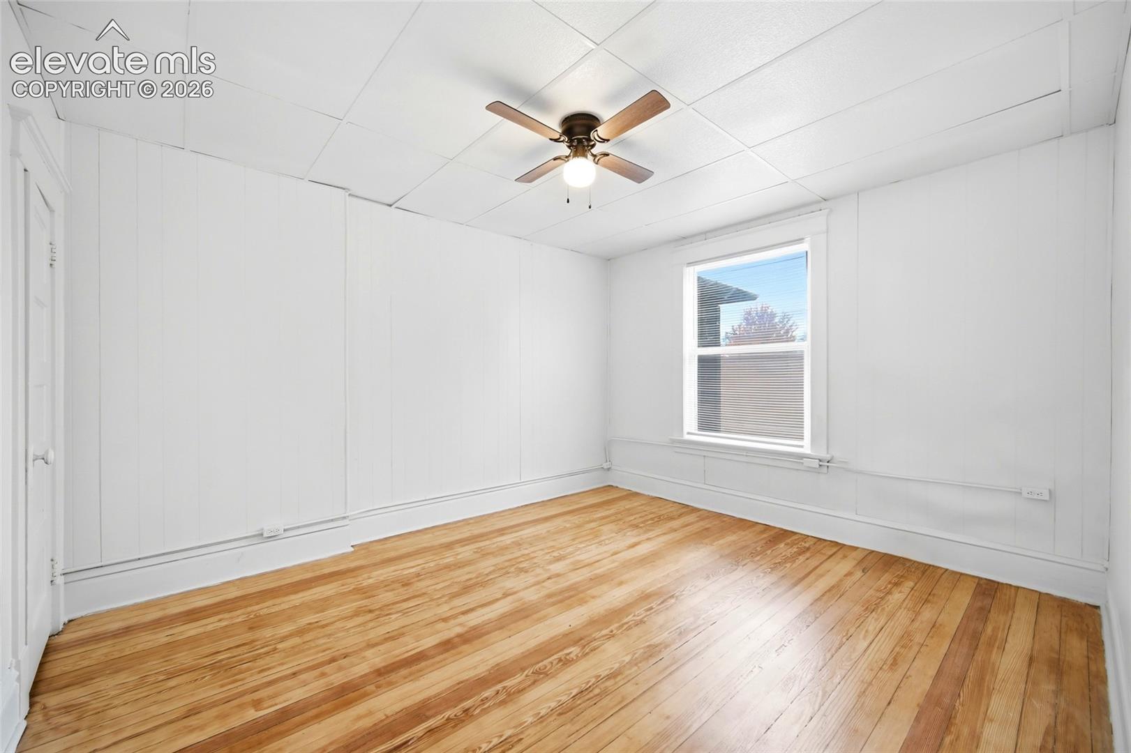 Interior room featuring wood-finish flooring, white panel walls, a tile ceiling, and a multi-blade ceiling fan with integrated lighting