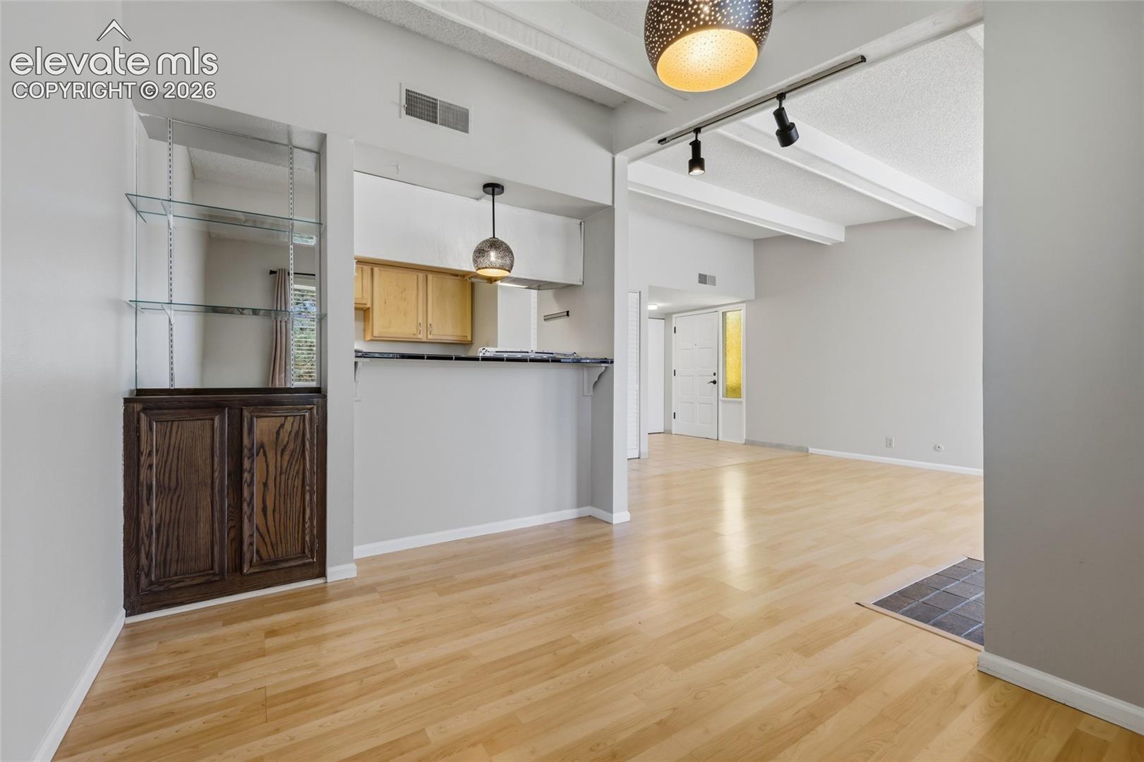 Wood-finish flooring, built-in lower cabinetry with overhead glass shelving, breakfast bar with tile countertop, vaulted ceiling with exposed beams, and decorative pendant lighting
