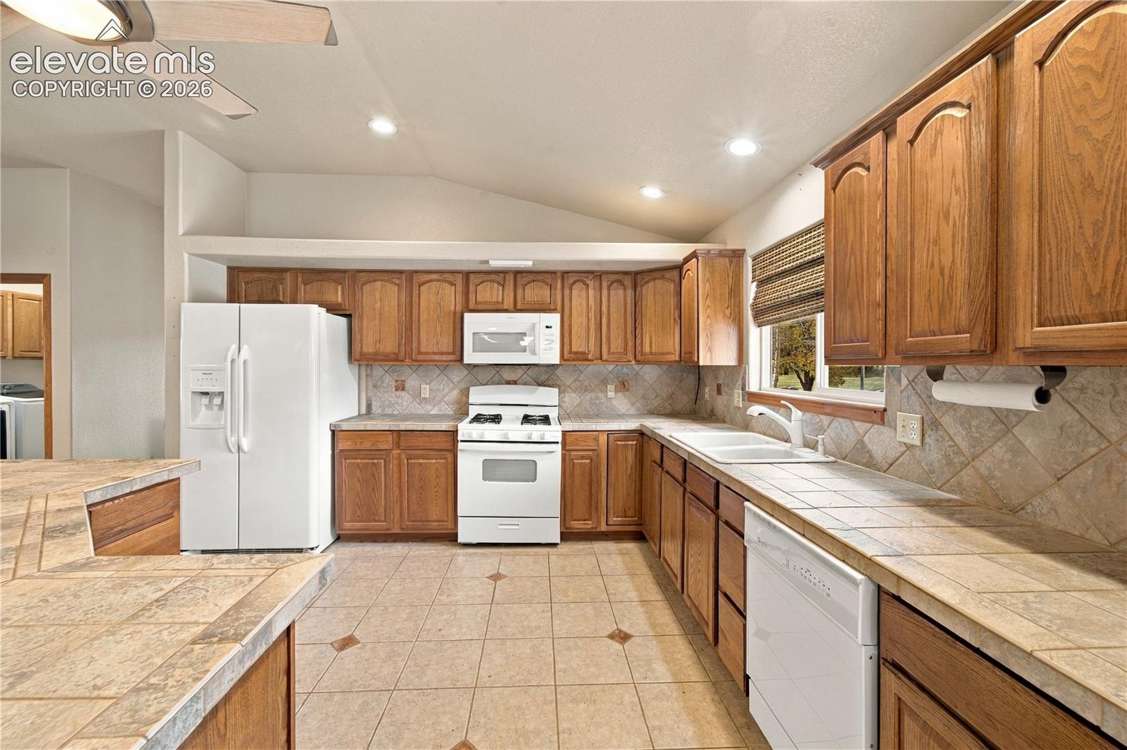 Kitchen featuring natural wood cabinetry, tile countertops with matching backsplash, ceramic tile flooring, and vaulted ceiling