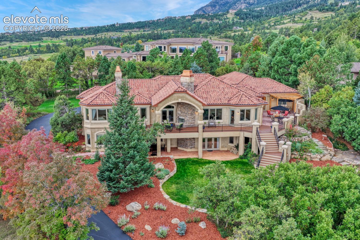 Stucco exterior featuring a clay tile roof, natural stone chimney, elevated deck with iron railing, and hardscaped staircase