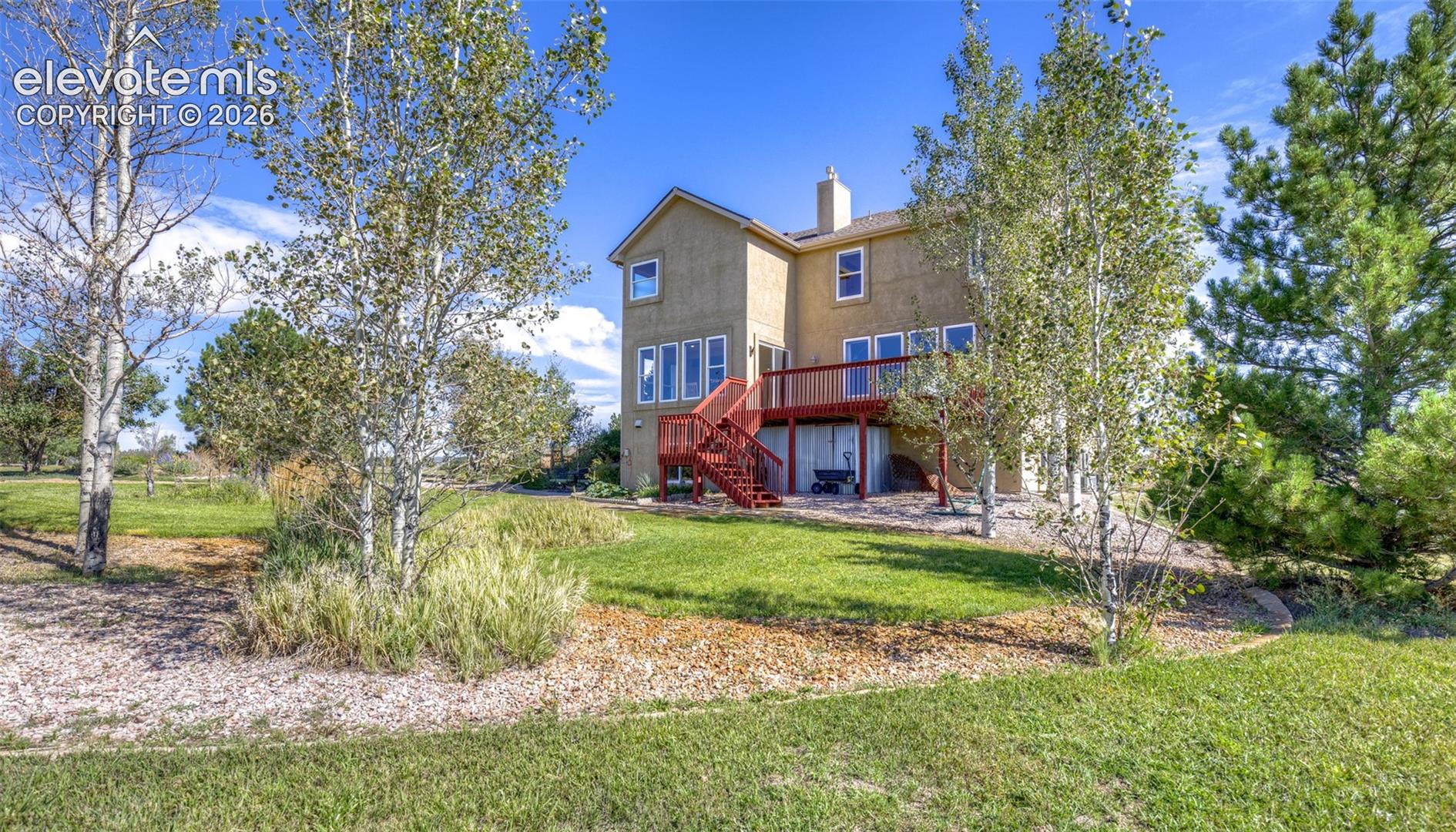 Stucco exterior with a multi-level red-stained wood deck, featuring multiple windows, a chimney, and surrounding mature trees and landscaping