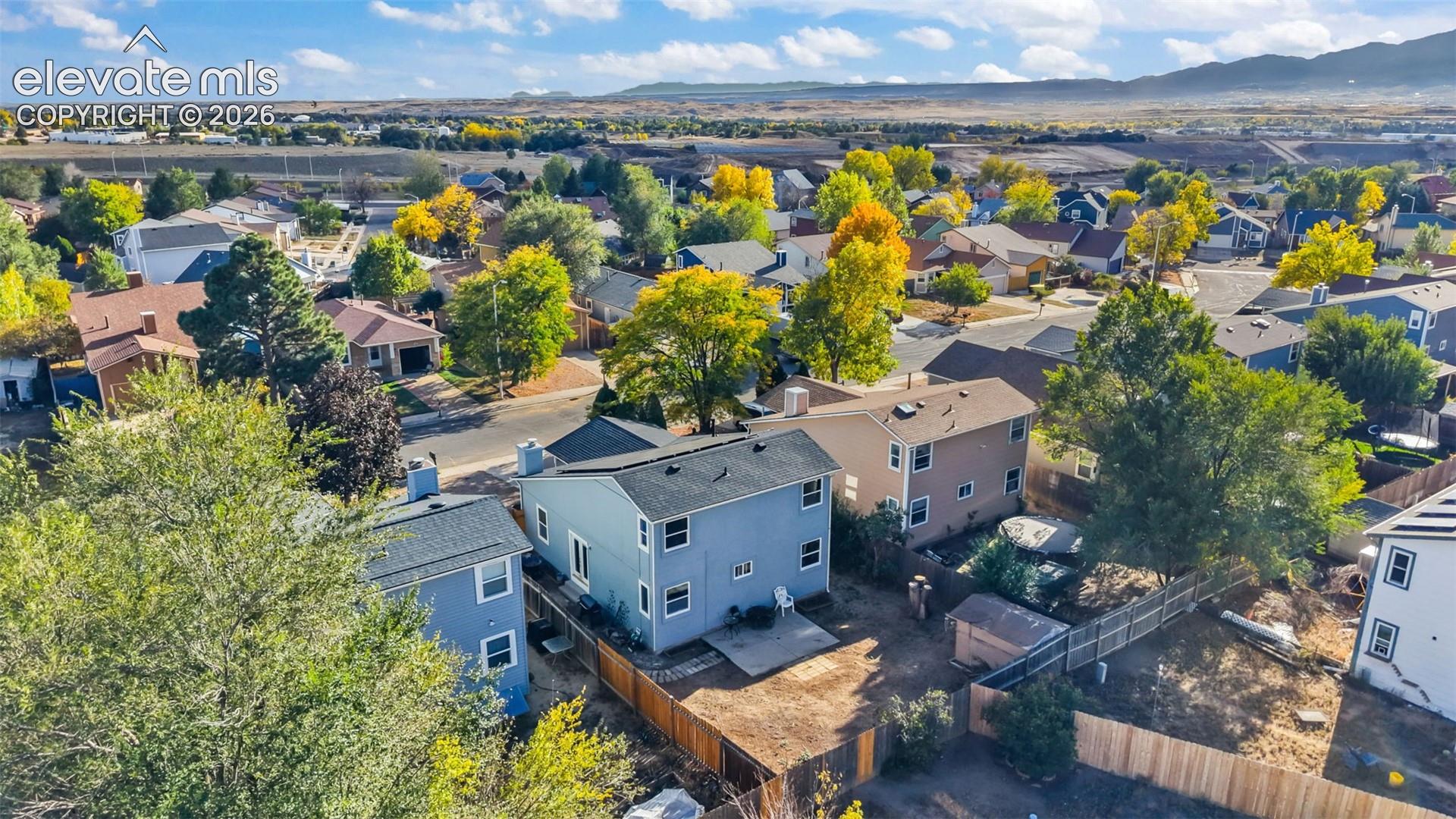 Aerial perspective featuring a two-story residence with a pitched shingle roof, a concrete patio, and a wood perimeter fence against a mountain backdrop