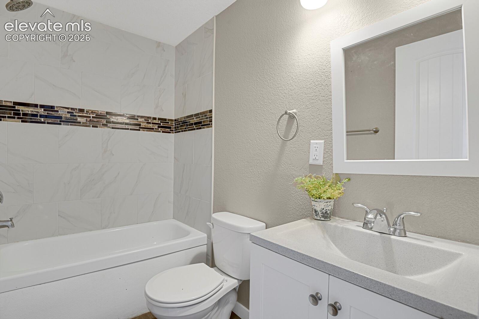 Bathroom featuring a tub with stone-finish tile surround, a glass and metal mosaic accent border, a white vanity with a composite countertop, and a framed wall mirror