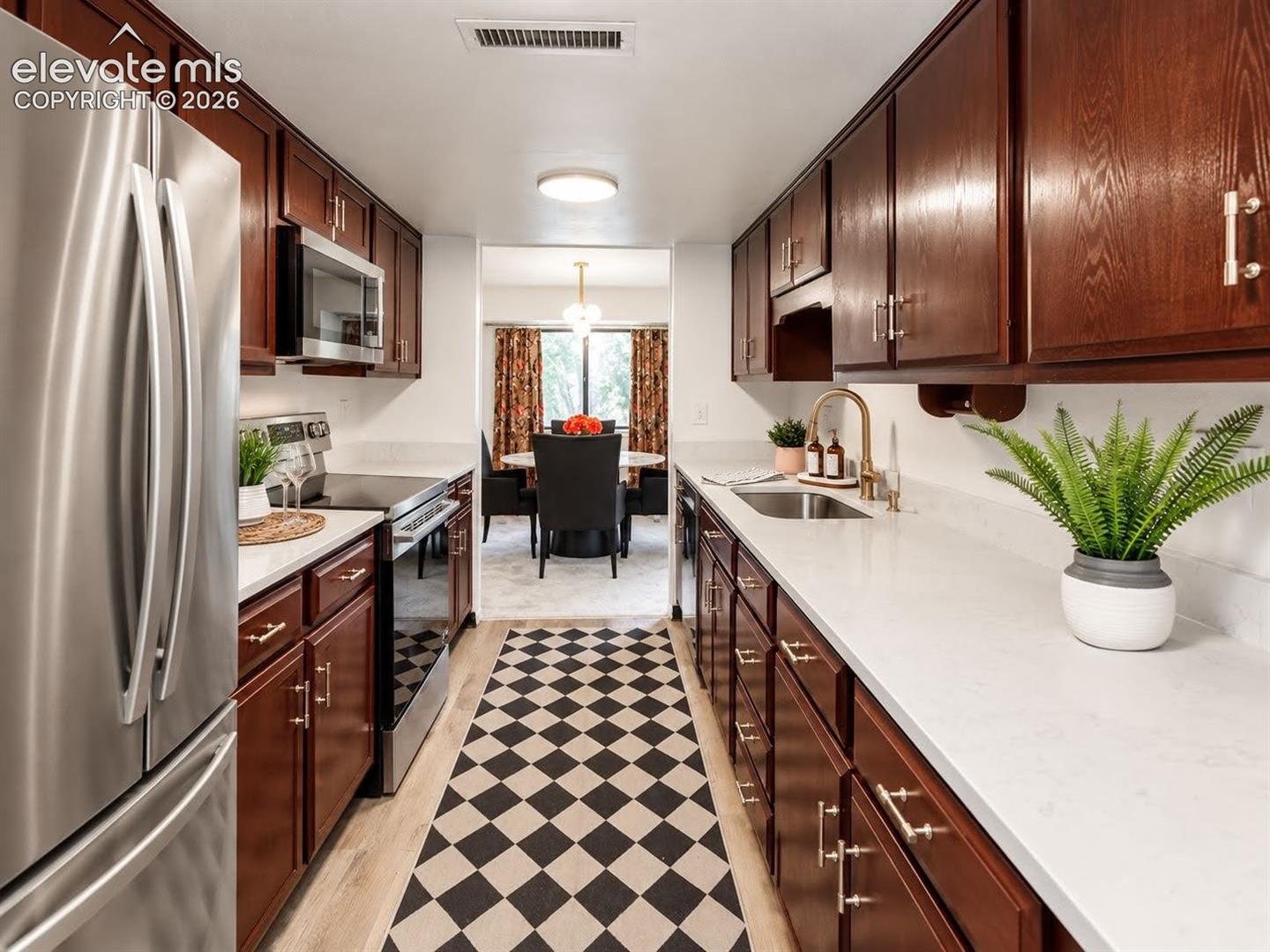 Galley kitchen featuring rich wood-toned cabinetry, light-colored stone-finish countertops, stainless steel appliances, and a modern brass-finish faucet