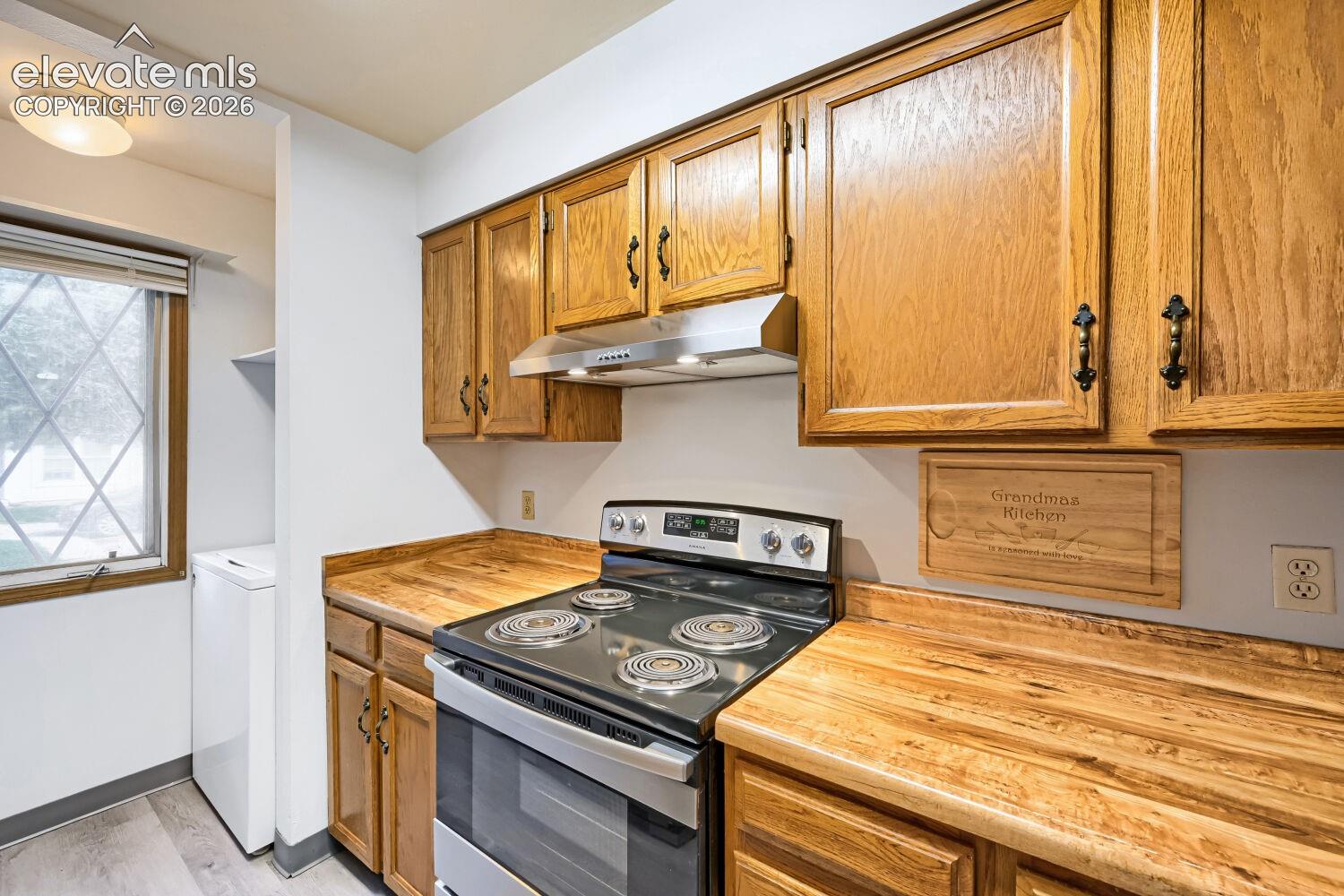 Wood-finish cabinetry with dark hardware, wood-finish countertops, stainless steel range with overhead ventilation, and a window with diamond-patterned grilles