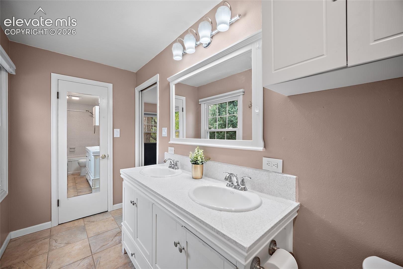 Double vanity with a stone-finish countertop, white cabinetry, chrome-finish dual-handle faucets, and a framed mirror featuring integrated four-bulb lighting