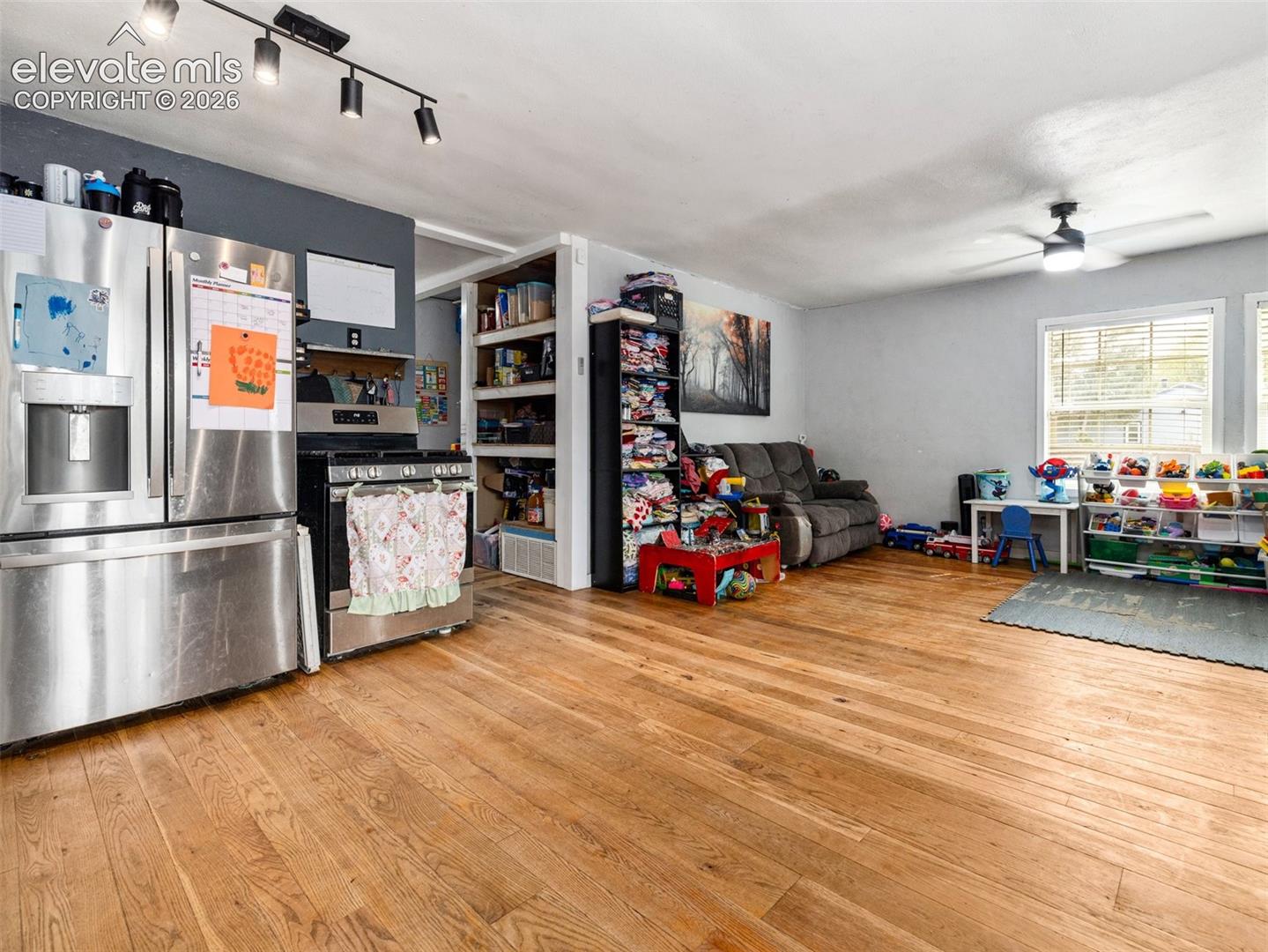 Expansive wood-finish flooring, stainless steel kitchen appliances, track lighting, ceiling fan, and large windows with blinds