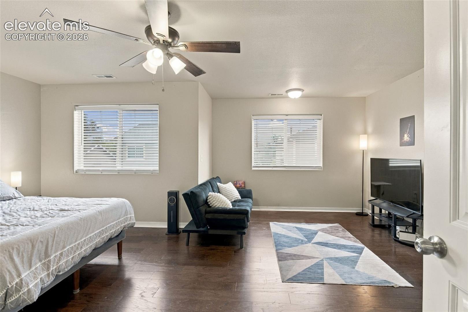 Spacious room featuring dark wood-finish flooring, dual windows with horizontal blinds, and a multi-light ceiling fan