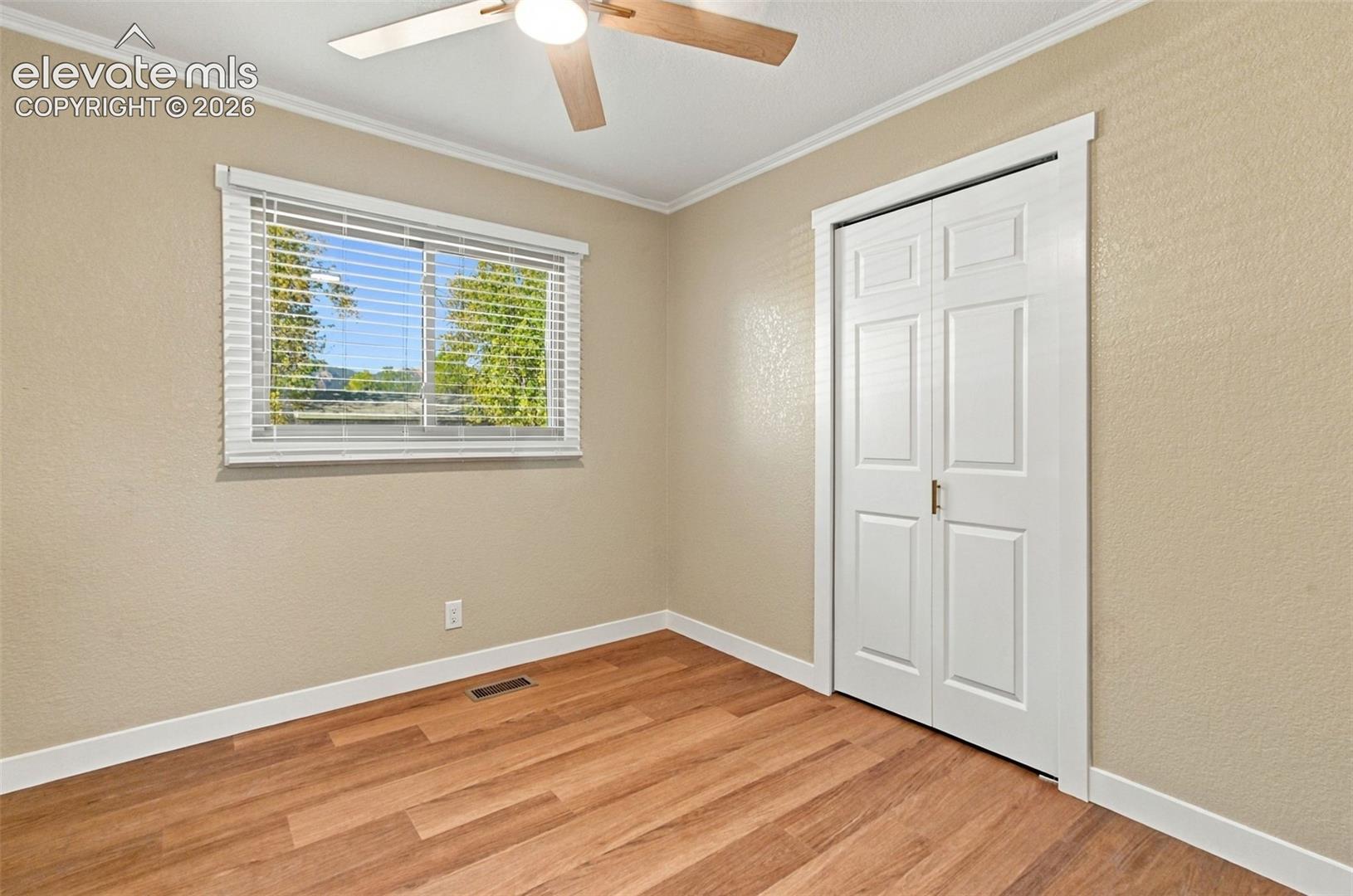Wood-finish flooring, white bi-fold closet doors, horizontal blind window covering, and ceiling fan with integrated lighting
