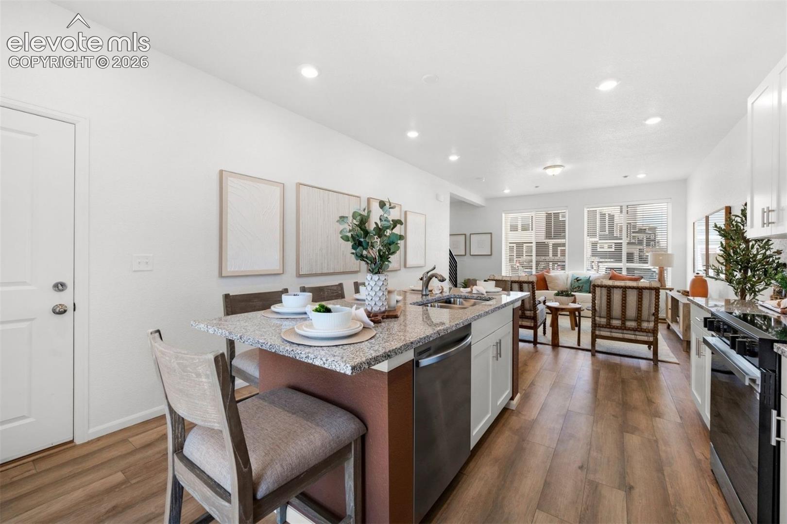 Open concept living space featuring wood-finish flooring throughout, a kitchen island with stone-look countertop, integrated stainless steel dishwasher, and white cabinetry
*photos of a model home*