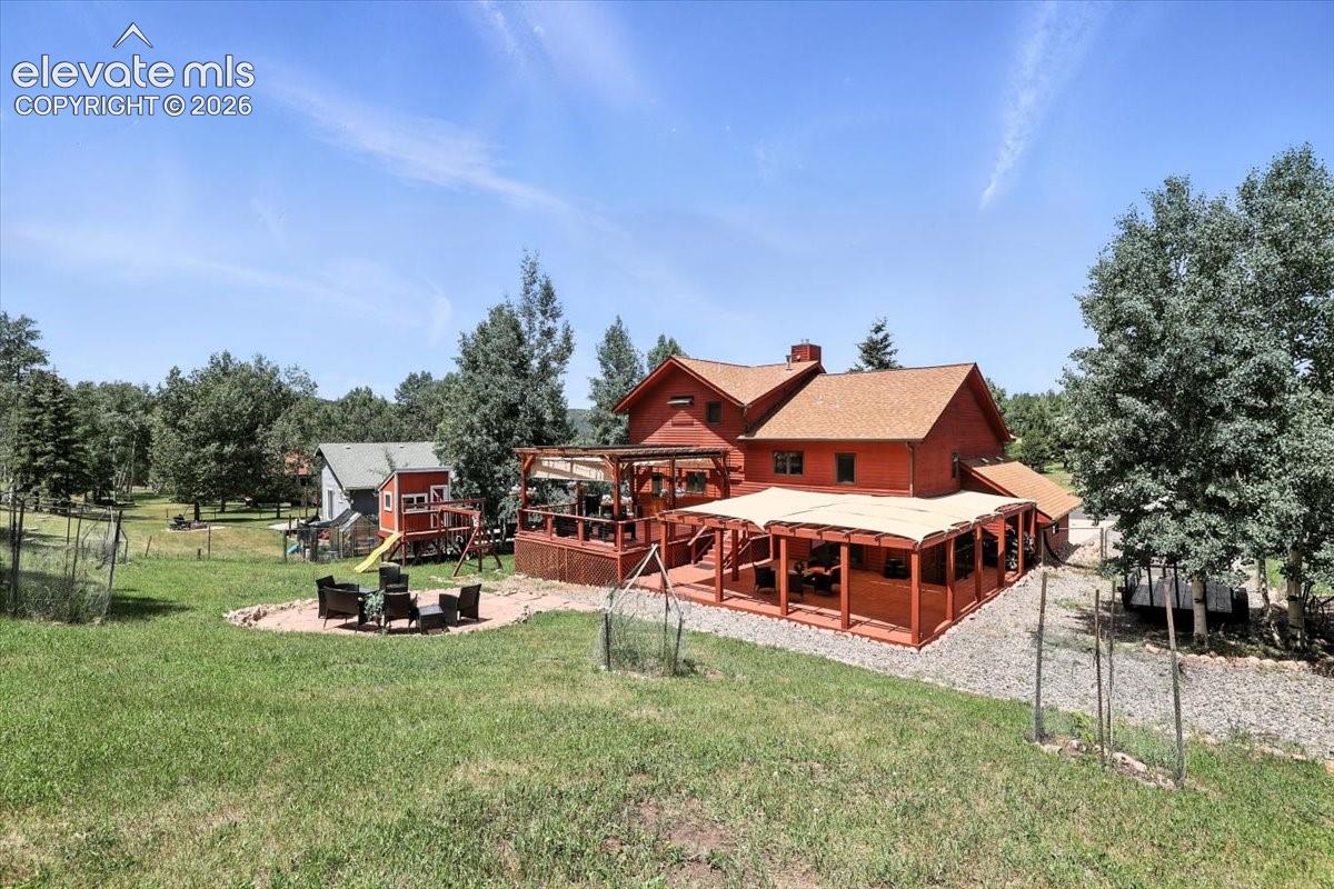 Red exterior residence featuring a covered wooden deck, pergola, paved stone patio, and expansive landscaped grounds with mature trees