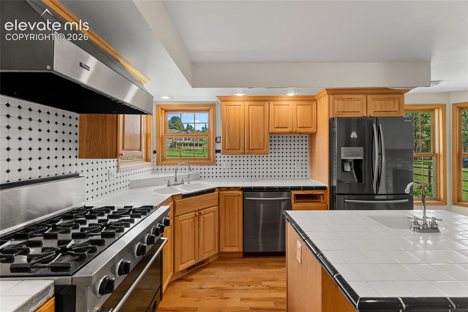 Kitchen featuring wood cabinetry, stainless steel range with hood, black appliances, tile countertops, and a tile backsplash