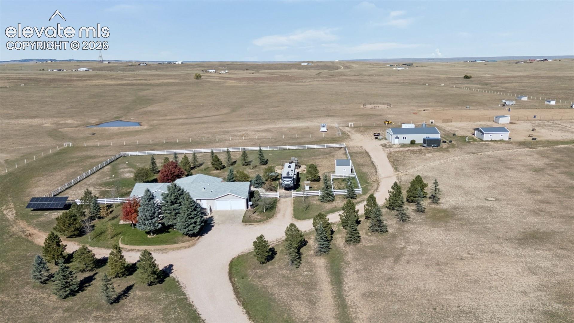 Aerial perspective of rural property featuring a primary residence with attached garage, ground-mounted solar array, detached outbuildings, and extensive perimeter fencing