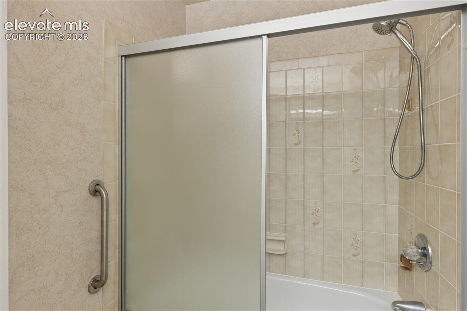 Tub and shower combination featuring a sliding frosted glass enclosure, light-toned ceramic tile surround, integrated metal grab bar, and wall-mounted shower head