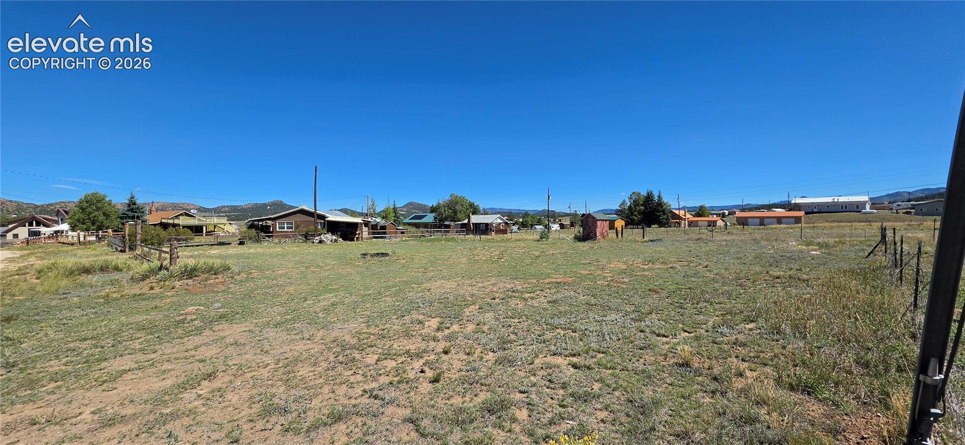 Expansive open field with natural ground cover, partial fencing, and distant residential structures